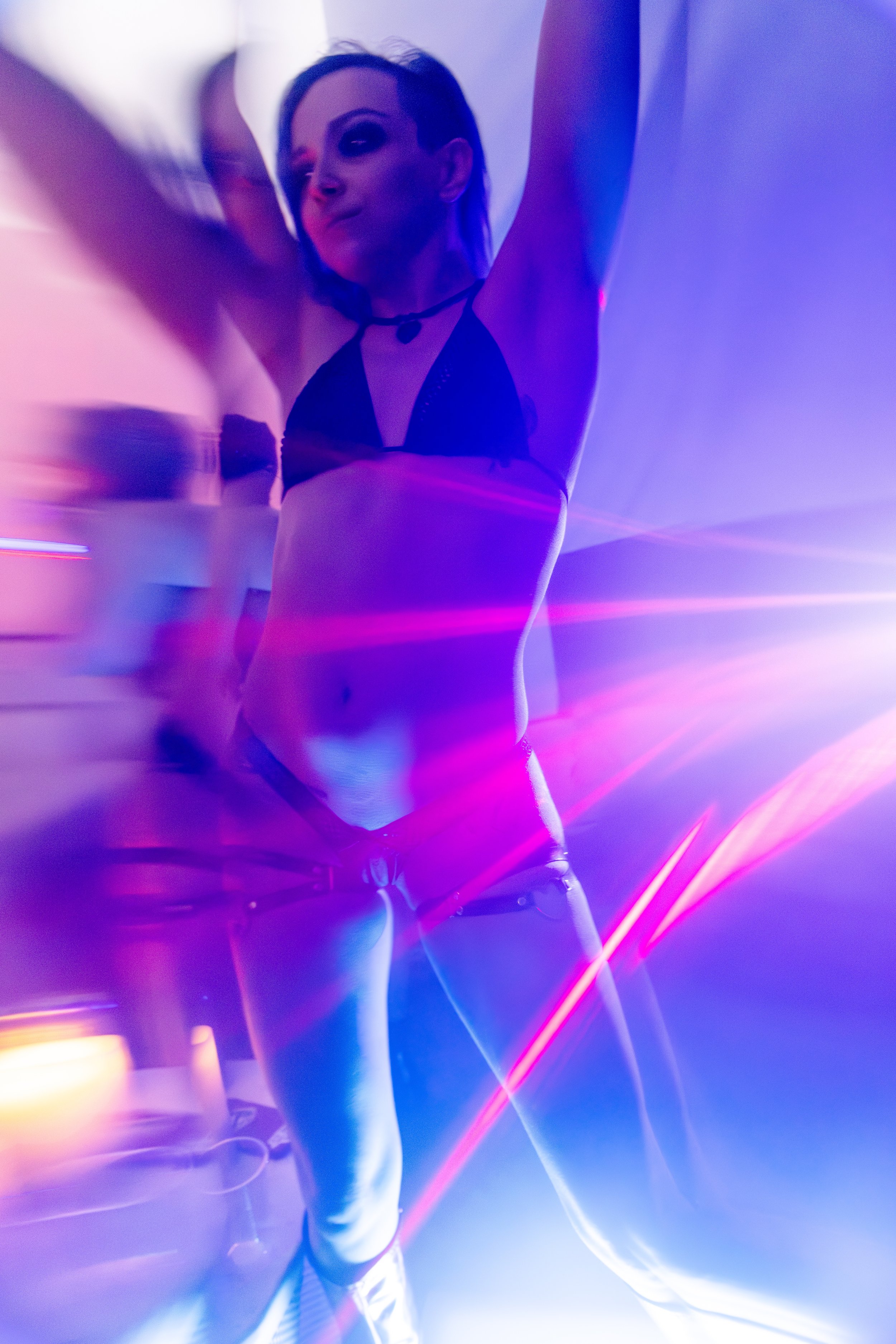 A woman with dark hair and makeup dancing in a club with colorful, vibrant lighting and laser effects.