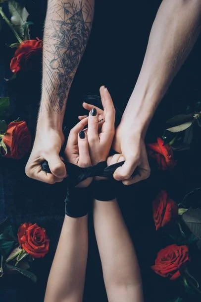 A person with tattoos on their arm and hand is tying the wrists of another person with black ribbon, surrounded by red roses.