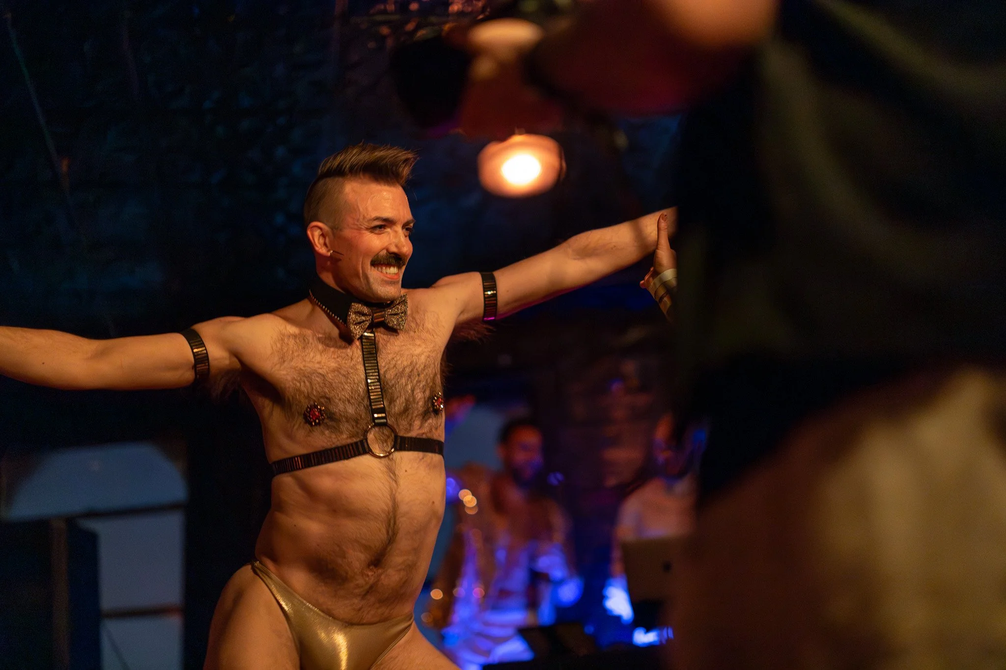 A man with a mustache and styled hair, wearing gold underwear, a bow tie, and various harnesses, is smiling with arms outstretched on a stage or in a performance setting.