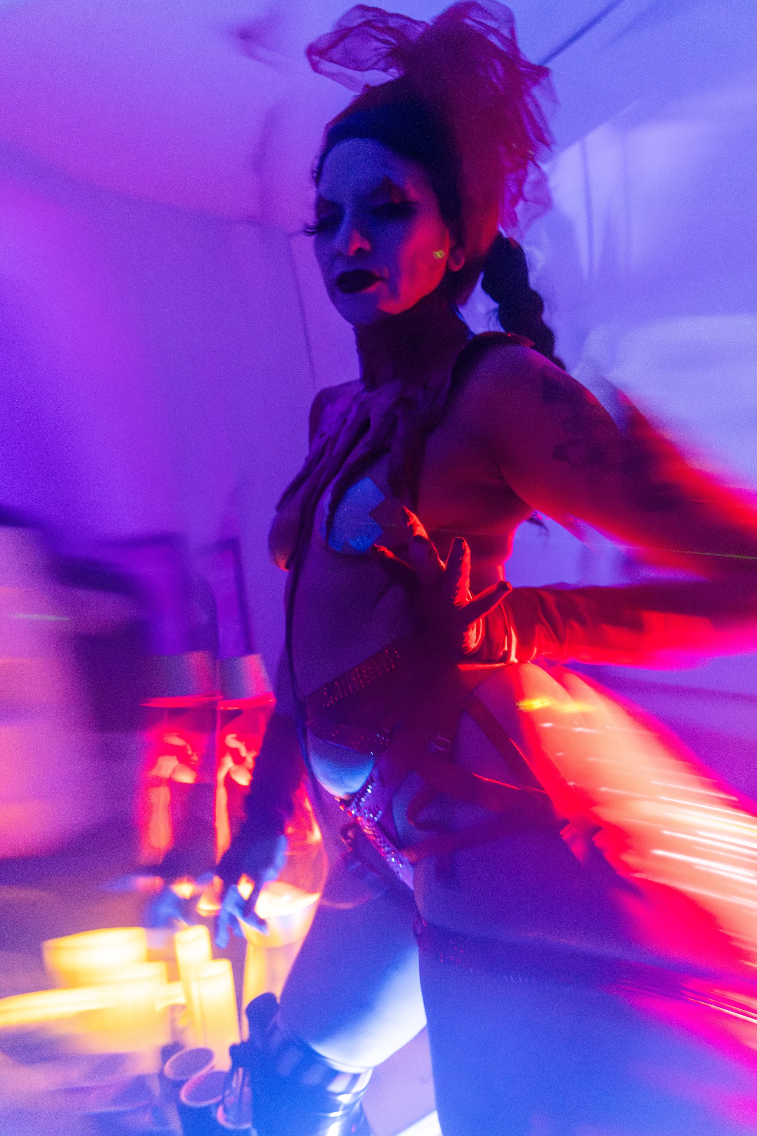 A woman at a performative event dressed in punk style with purple and red lighting, wearing a headpiece, gloves, and metallic clothing.