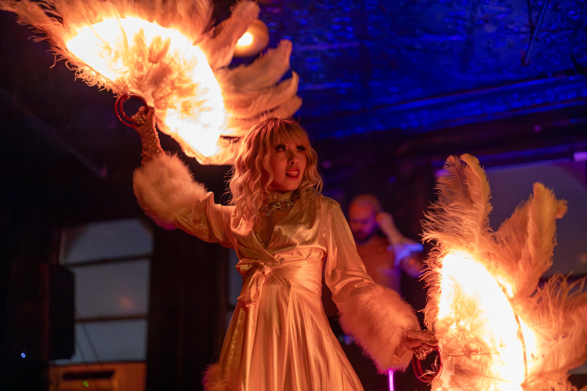 A woman performs a fire dance with flaming fans, wearing a satin gold dress with fluffy cuffs, in a dimly lit venue with blue and purple lighting.