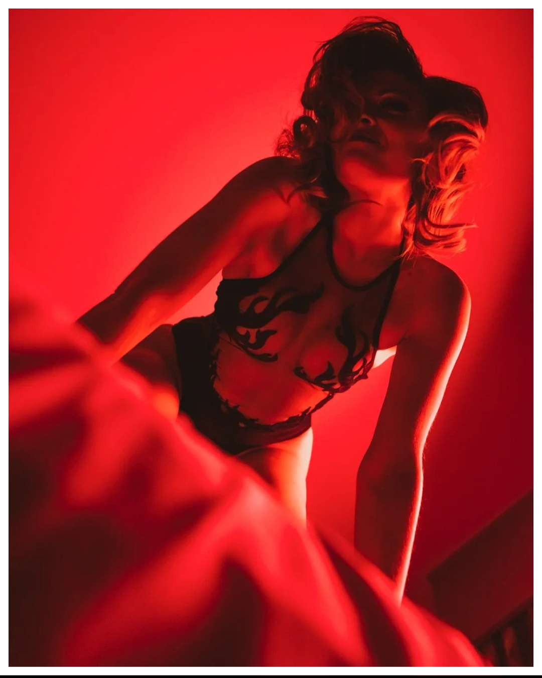 A woman in black lingerie with flame designs, illuminated by red lighting, leaning forward with her face partially obscured and her hair styled in loose curls.
