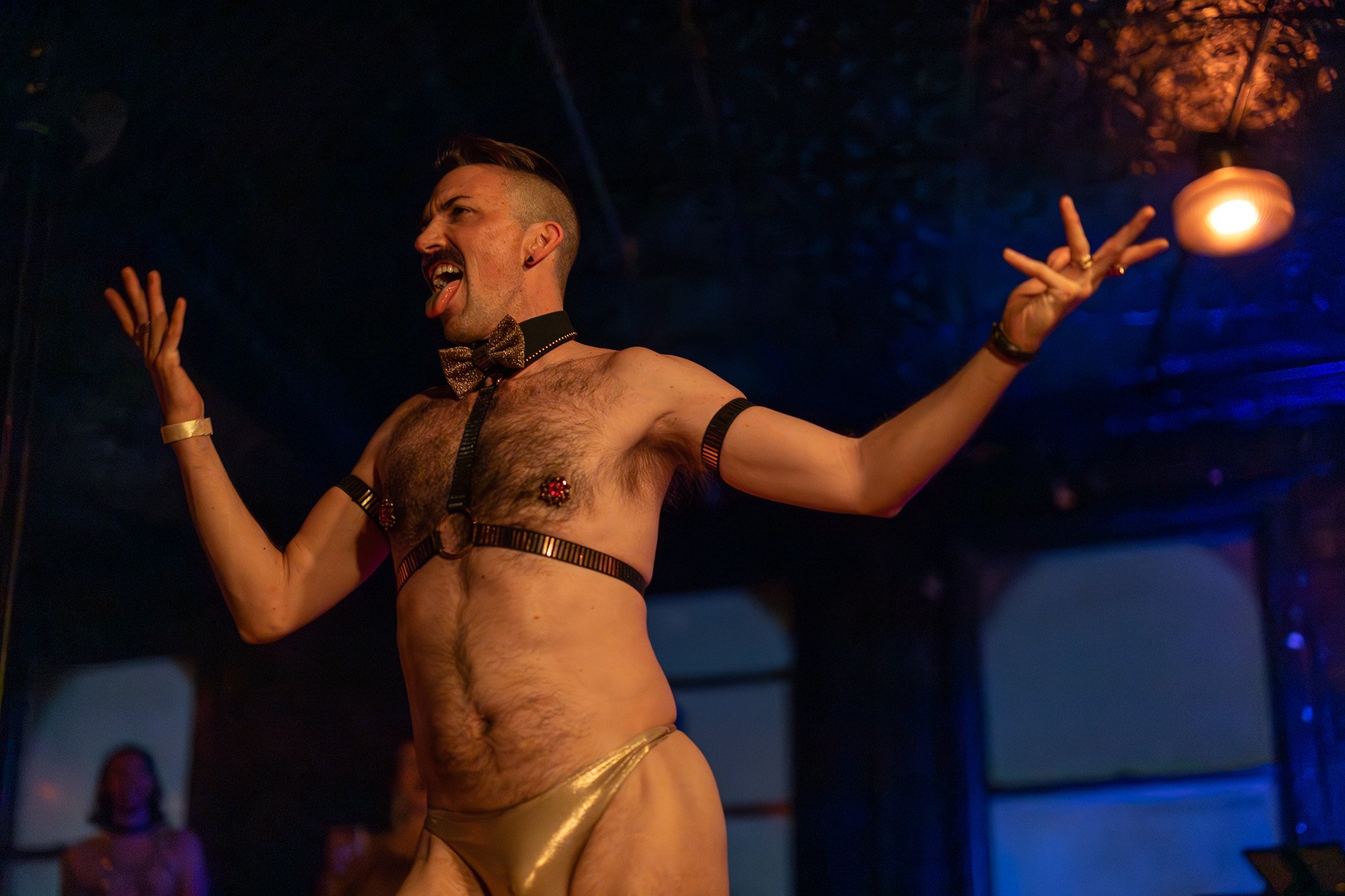 A man on stage wearing gold speedo, a bow tie, and harness with pasties, making a playful expression with tongue out and arms raised, in a dark room with colorful lighting.