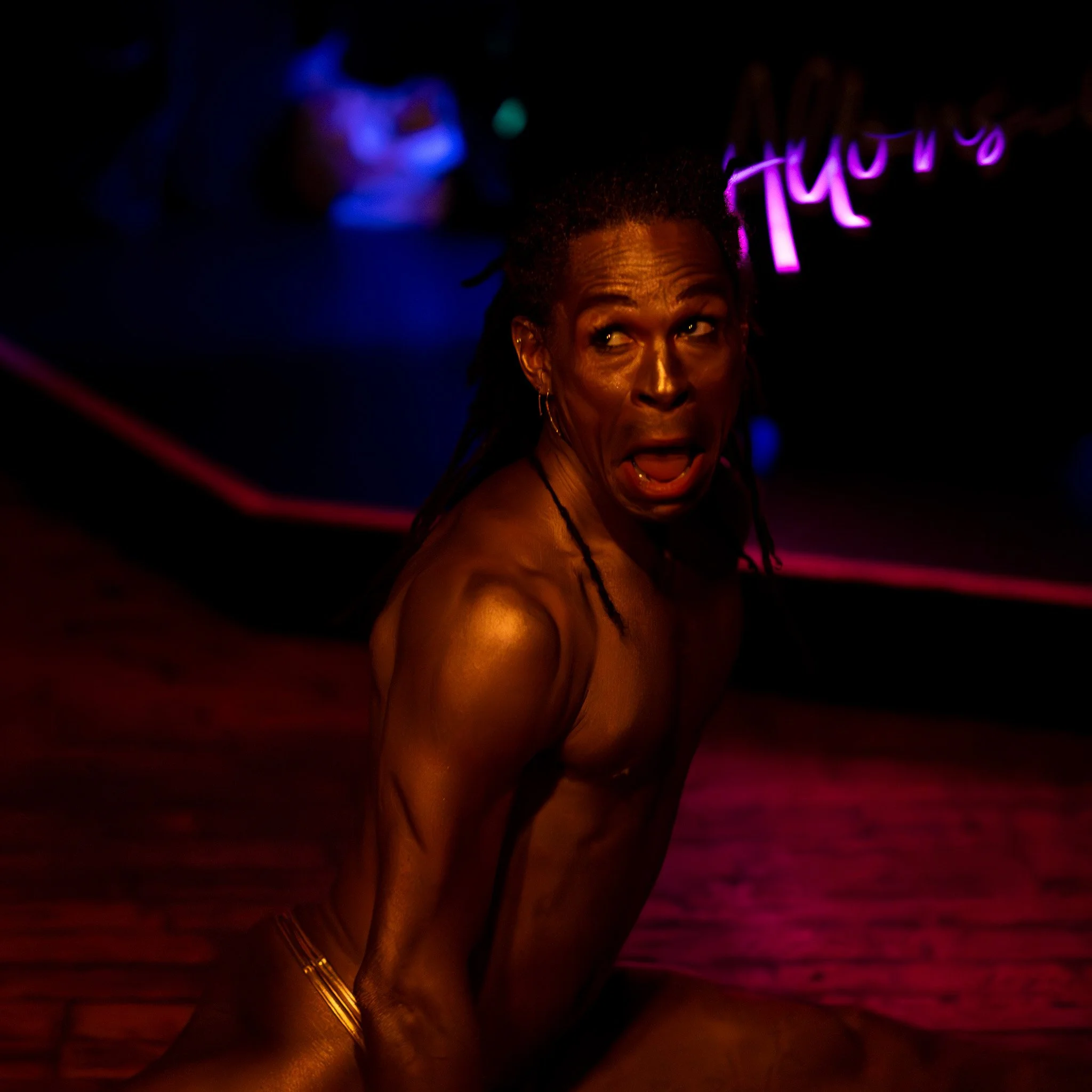 A topless woman with dreadlocks performing a dance or theatrical act in a dark setting with neon purple and pink lights.