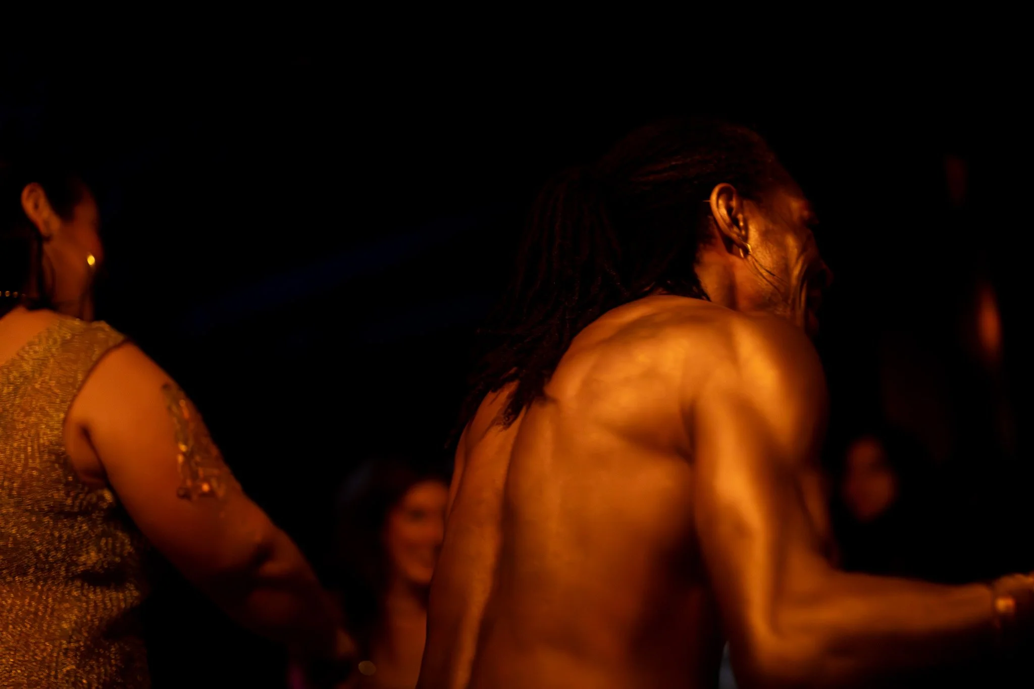 A shirtless man with dreadlocks smiling and looking to his right at a dimly lit event, with a woman in a gold dress and earrings nearby.