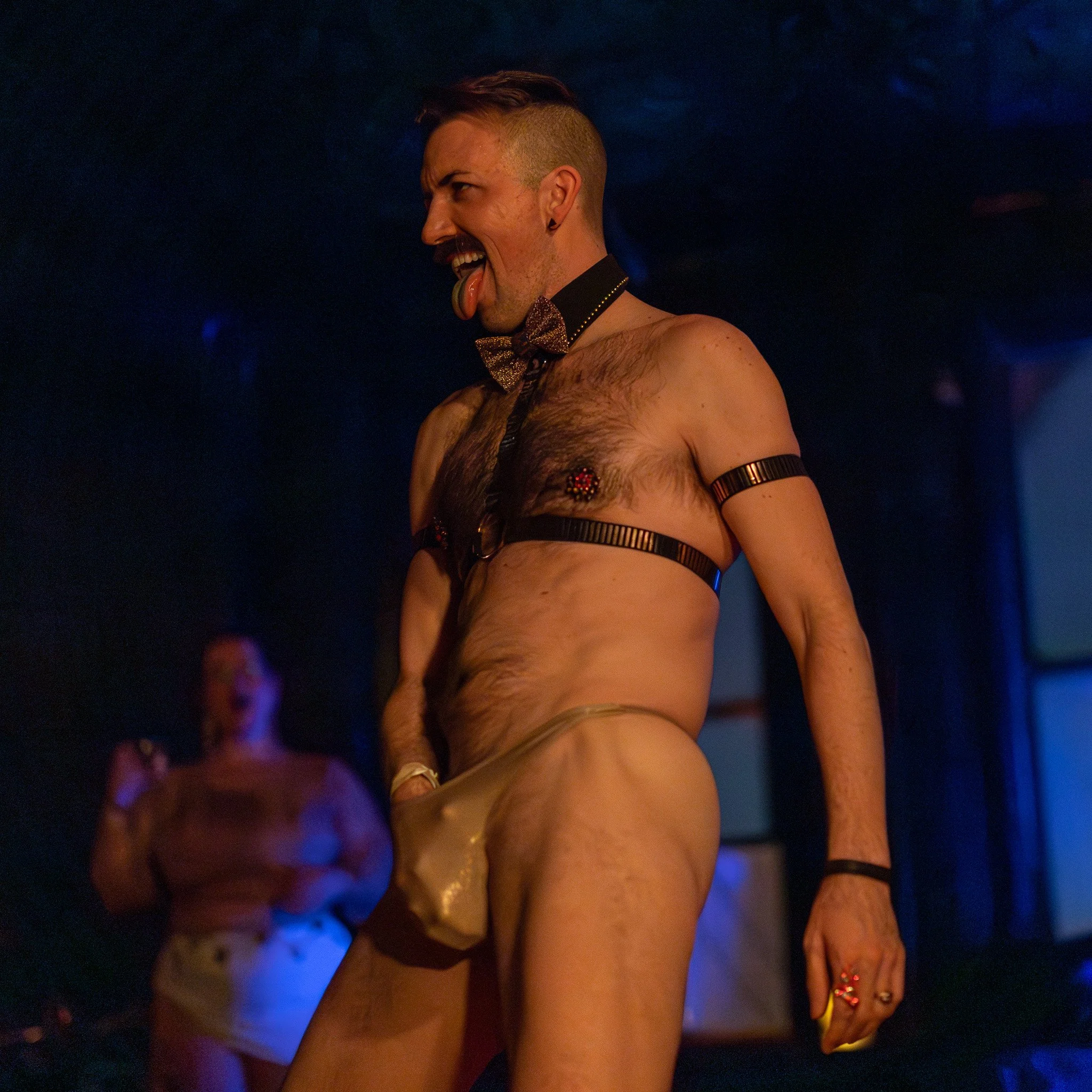 A man in a bowtie, harness, and leather accessories, sticking out his tongue and posing on stage, with a woman in the background.