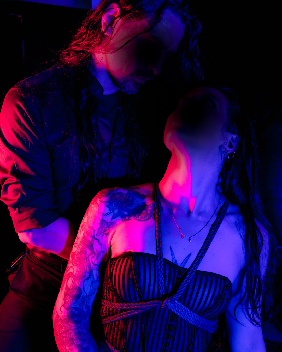 A man with long wavy hair and a beard leans over a woman with long dark hair and tattoos on her arm. The scene is lit with vibrant pink and blue neon lights, creating a dramatic and moody atmosphere.