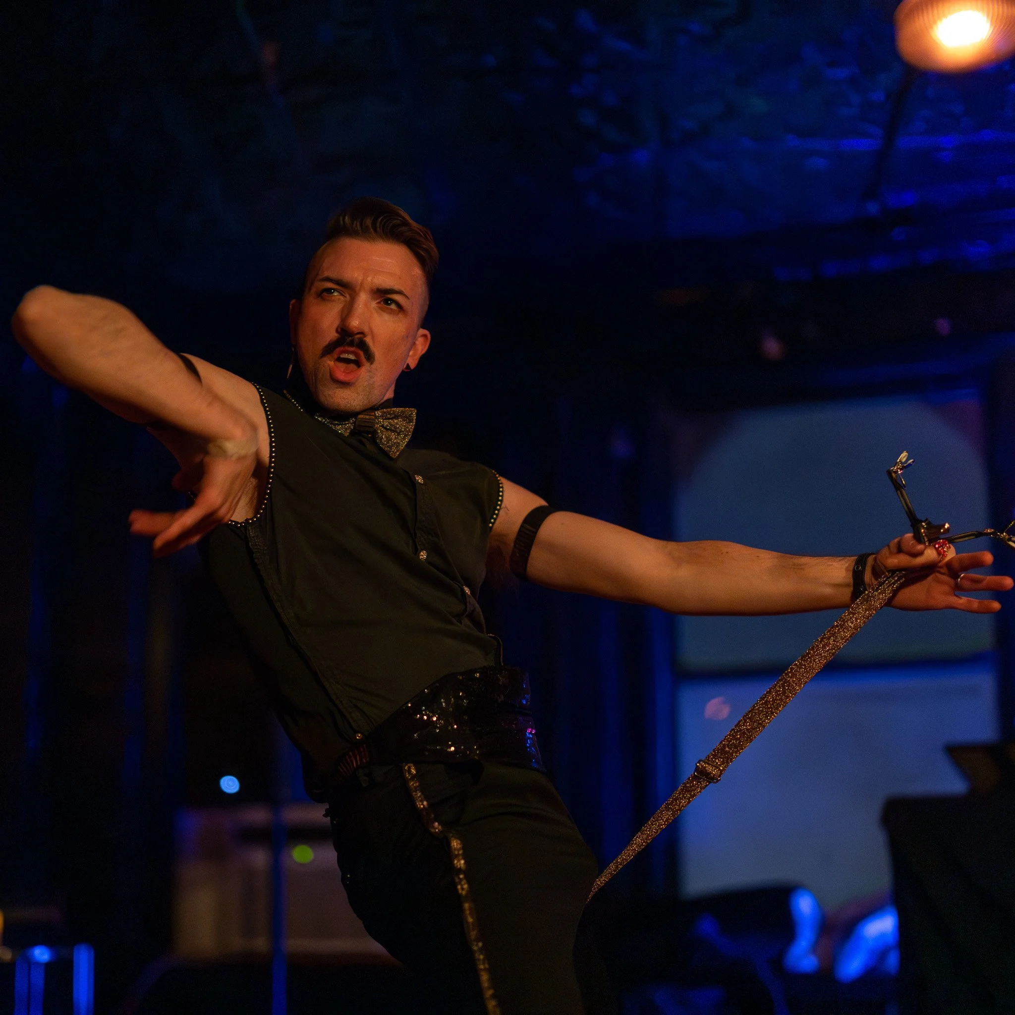 Performer with styled hair and mustache singing on stage, wearing black shirt with gold accents and holding a gold glittery microphone stand, illuminated by stage lights.