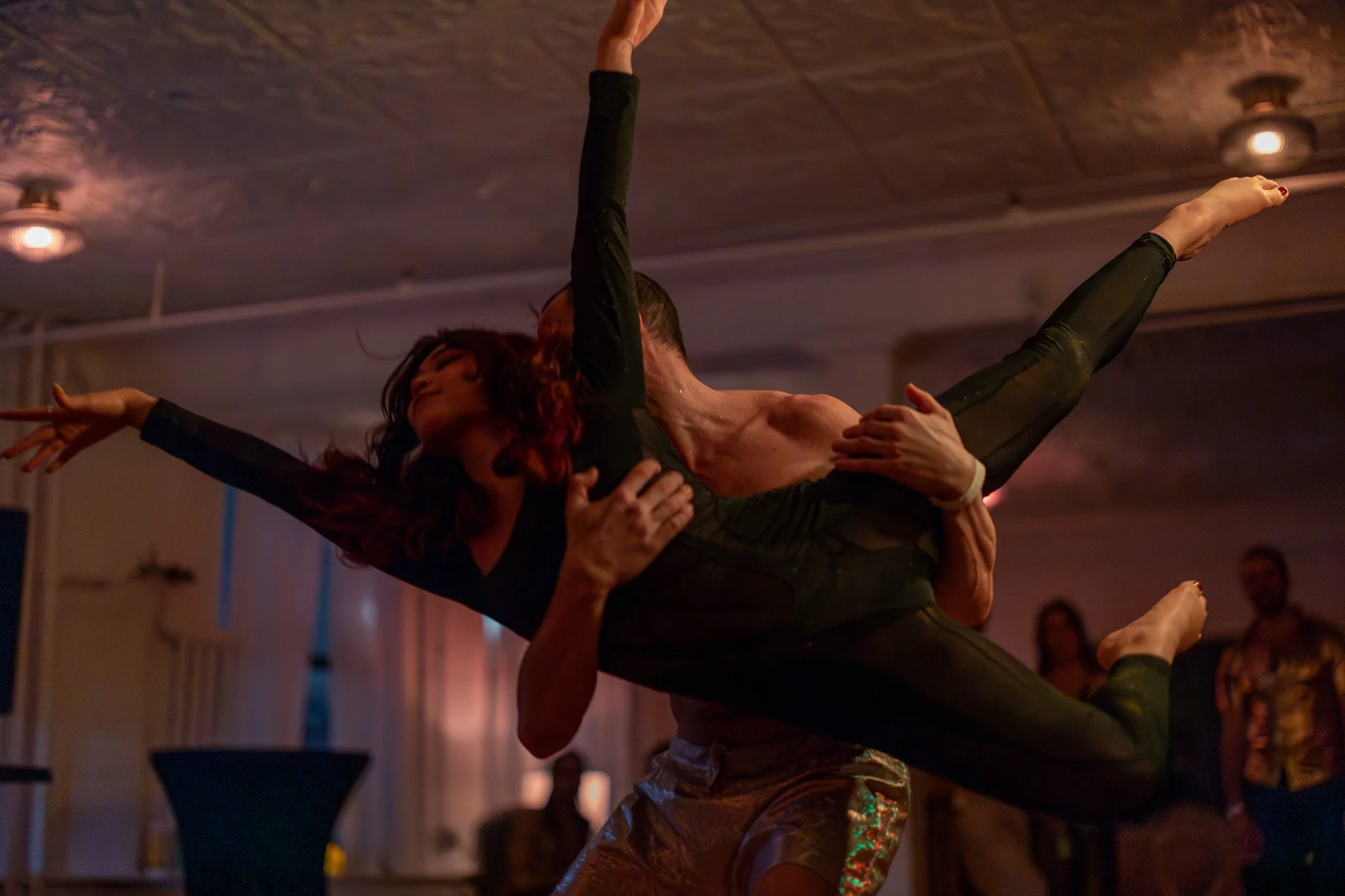 A male dancer lifts a female dancer during a performance in a dimly lit room with a textured ceiling, with onlookers in the background.