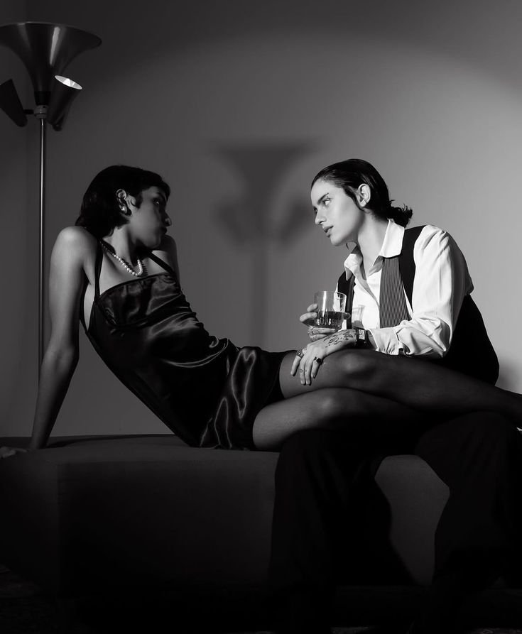 Black and white photo of two women in an intimate setting. One woman is sitting on a surface wearing a satin dress, and the other woman, dressed in a shirt and vest, is sitting close to her, holding a glass of wine and touching her leg.