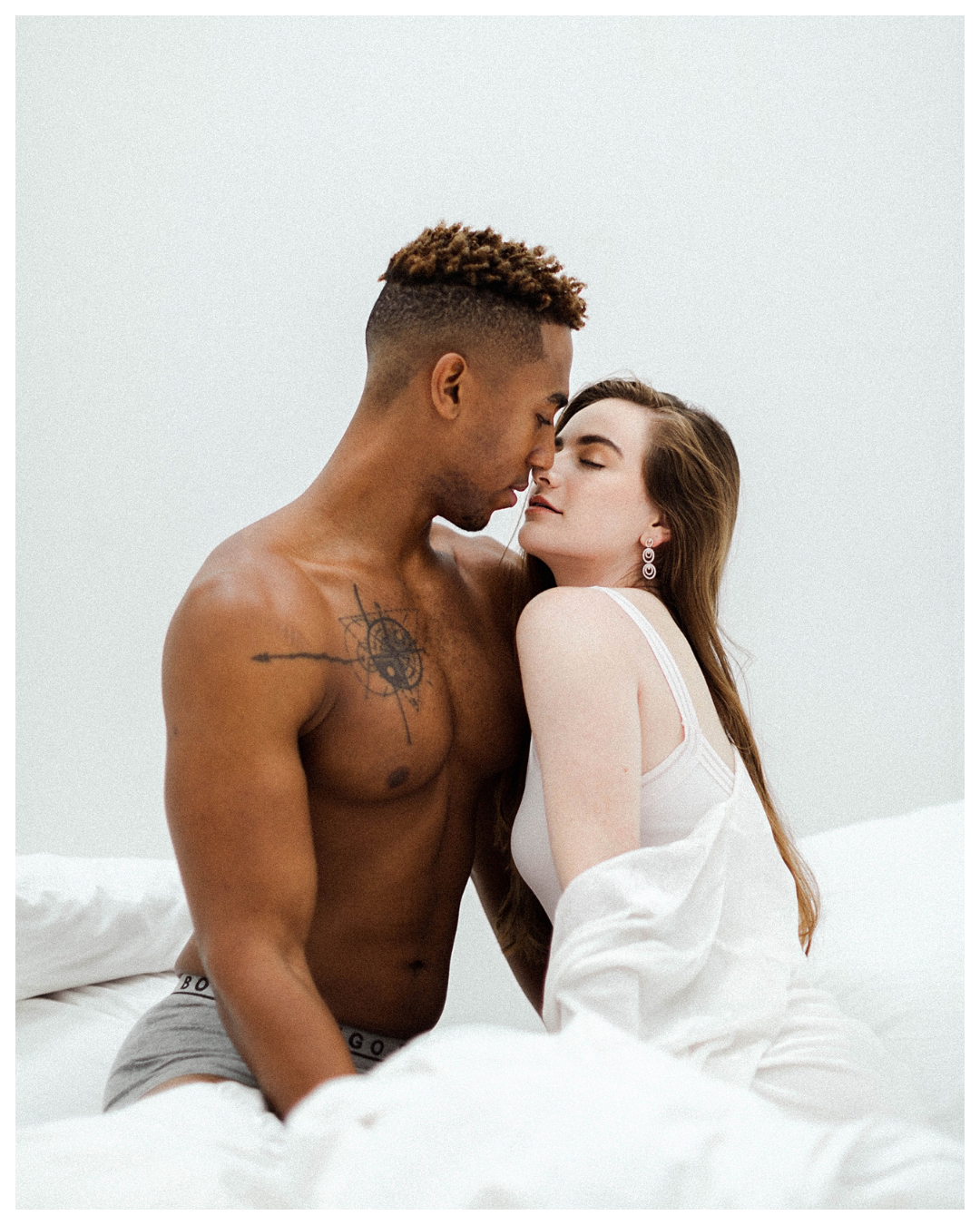 A shirtless man and a woman in white lingerie sitting closely on a bed with white sheets, touching foreheads and eyes closed.