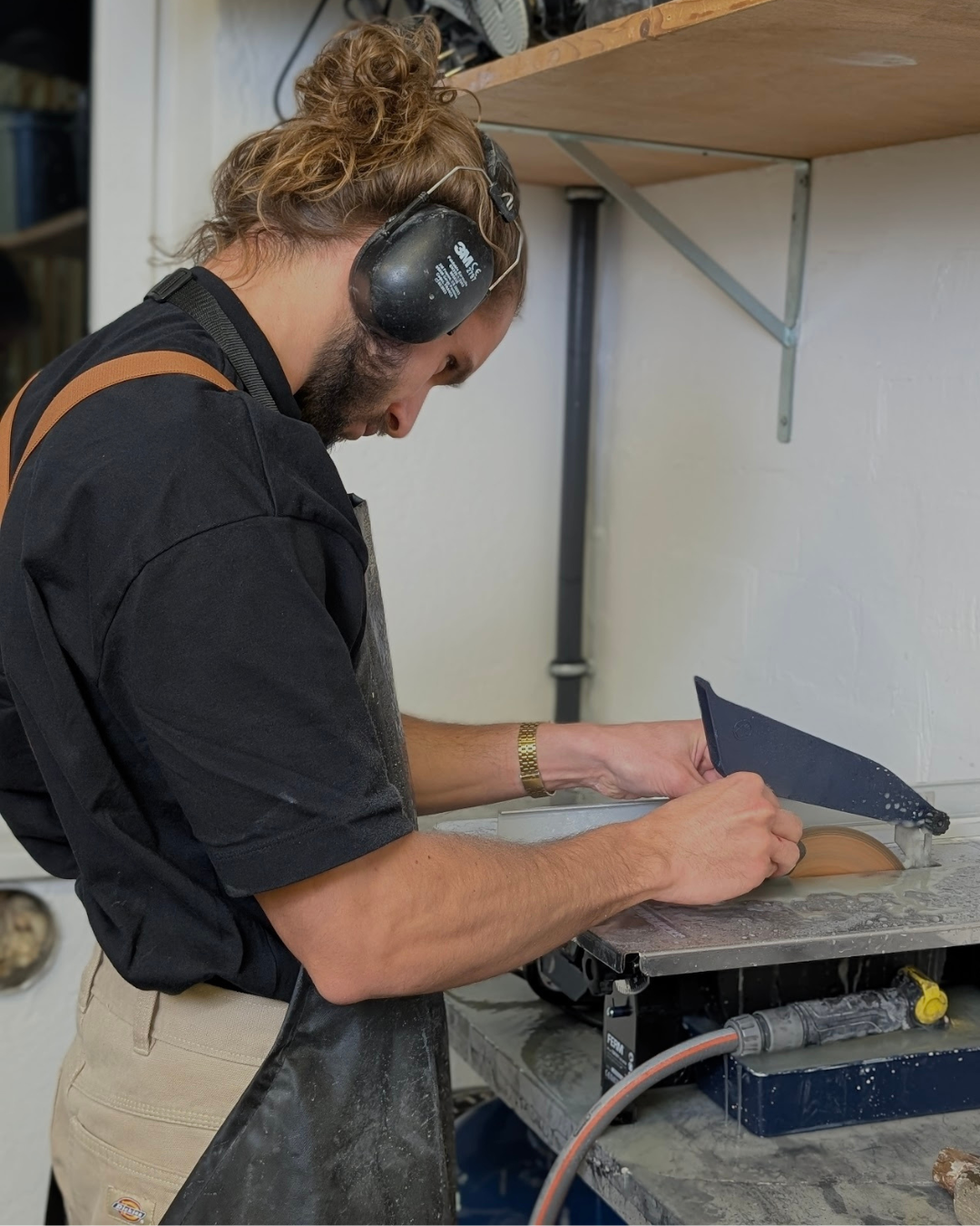 A man working in a workshop, wearing ear protection, an apron, and a black shirt, cutting a piece of wood with a circular saw on a workbench.