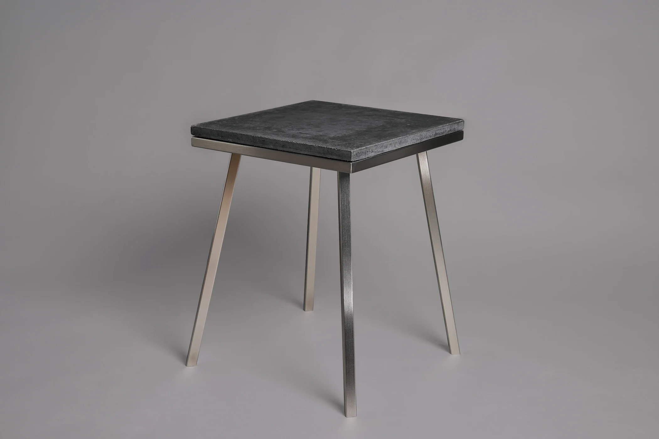 A modern stool with four angled metal legs and a flat, square, black stone seat against a plain gray background.