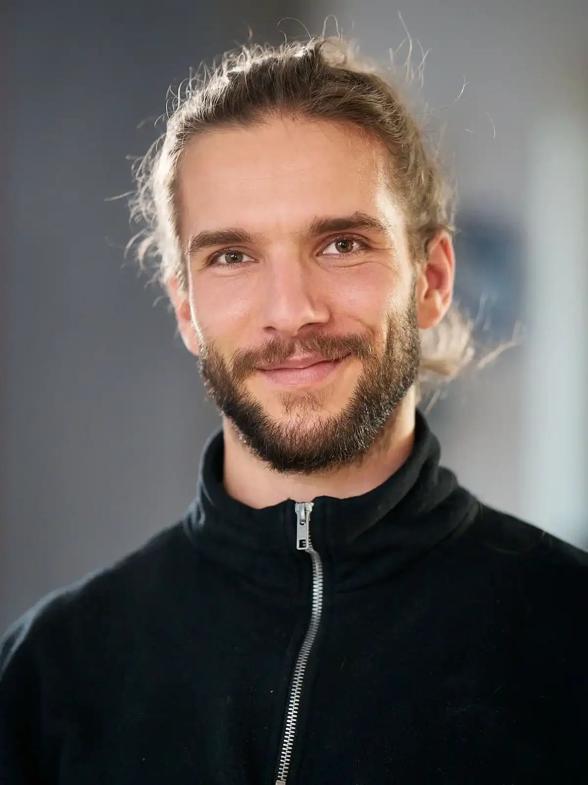 A smiling man with a beard and long hair tied back, wearing a black zip-up jacket, standing indoors with a blurred background.