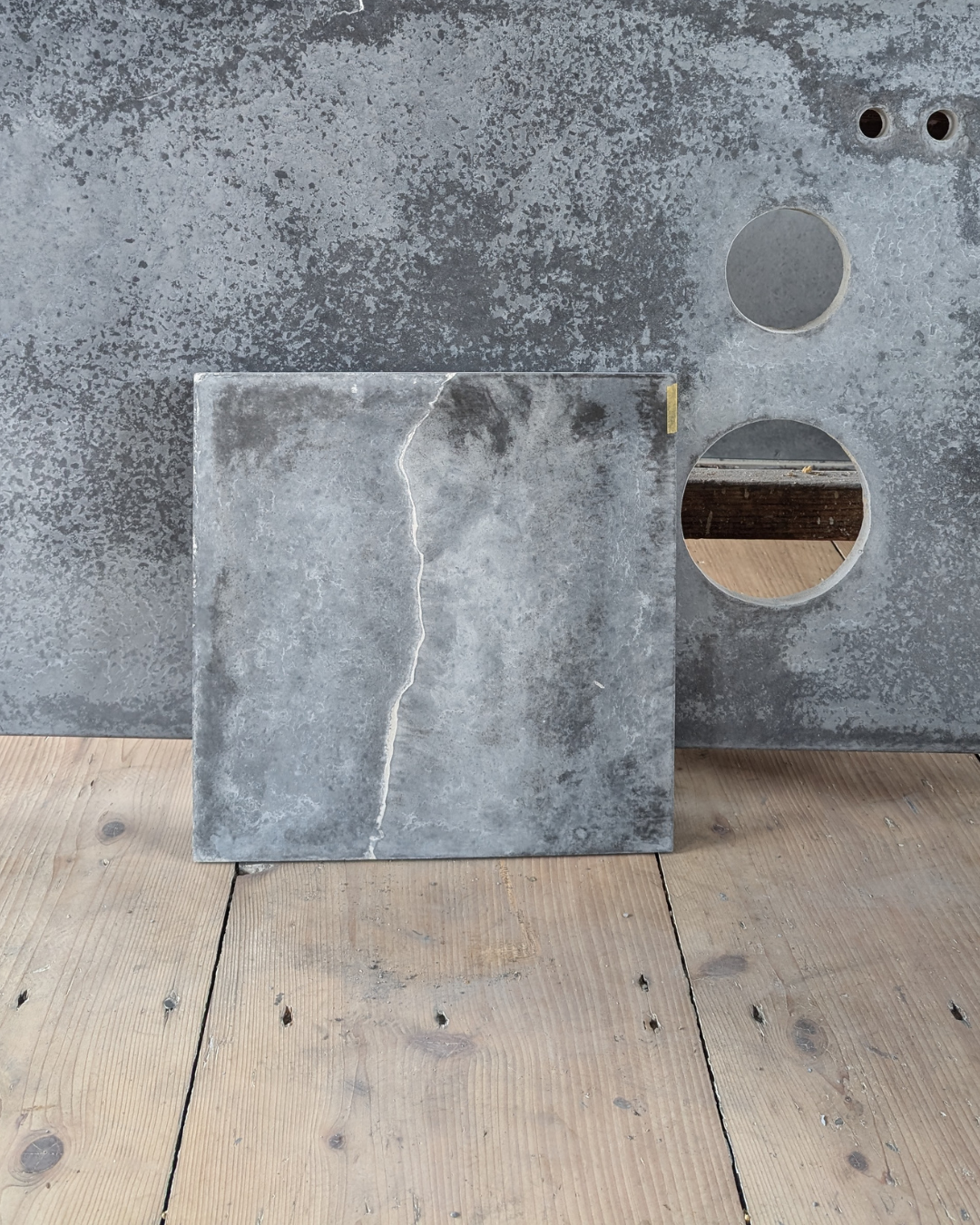 A close-up of a concrete square with a visible crack, leaning against a larger concrete surface with three circular holes. The surface and the square have a weathered, textured appearance, positioned on wooden floorboards.