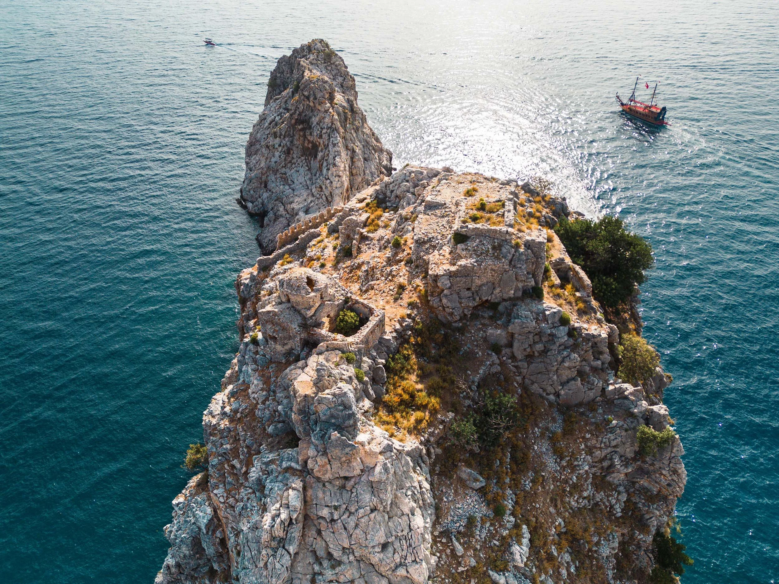 An aerial view of a rocky island with ancient roman church and a large solitary rock formation in the sea, with a boat nearby.