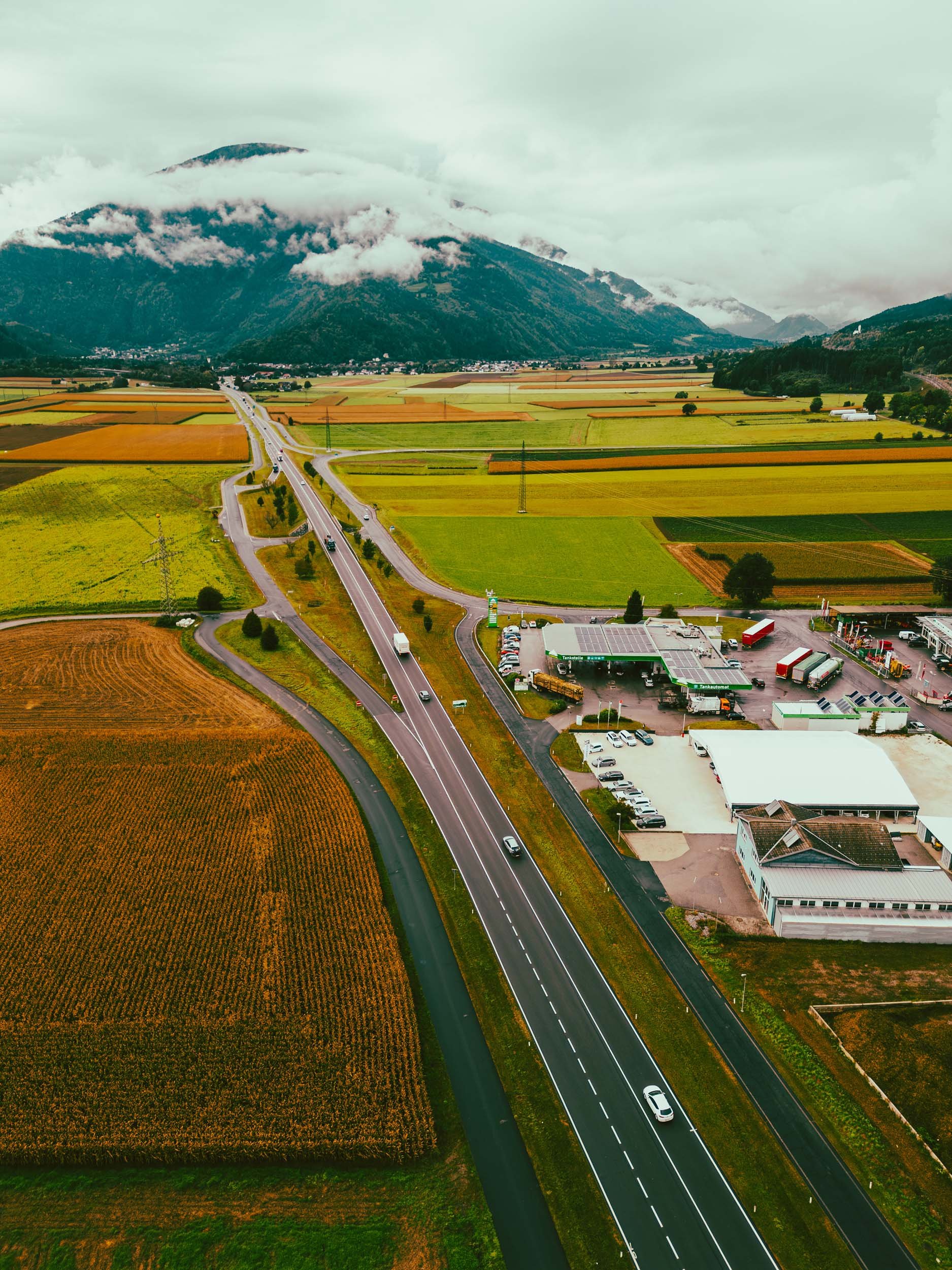 Aerial view of a rural area with a highway running through agricultural fields and a small commercial area, with mountains covered in clouds in the background.