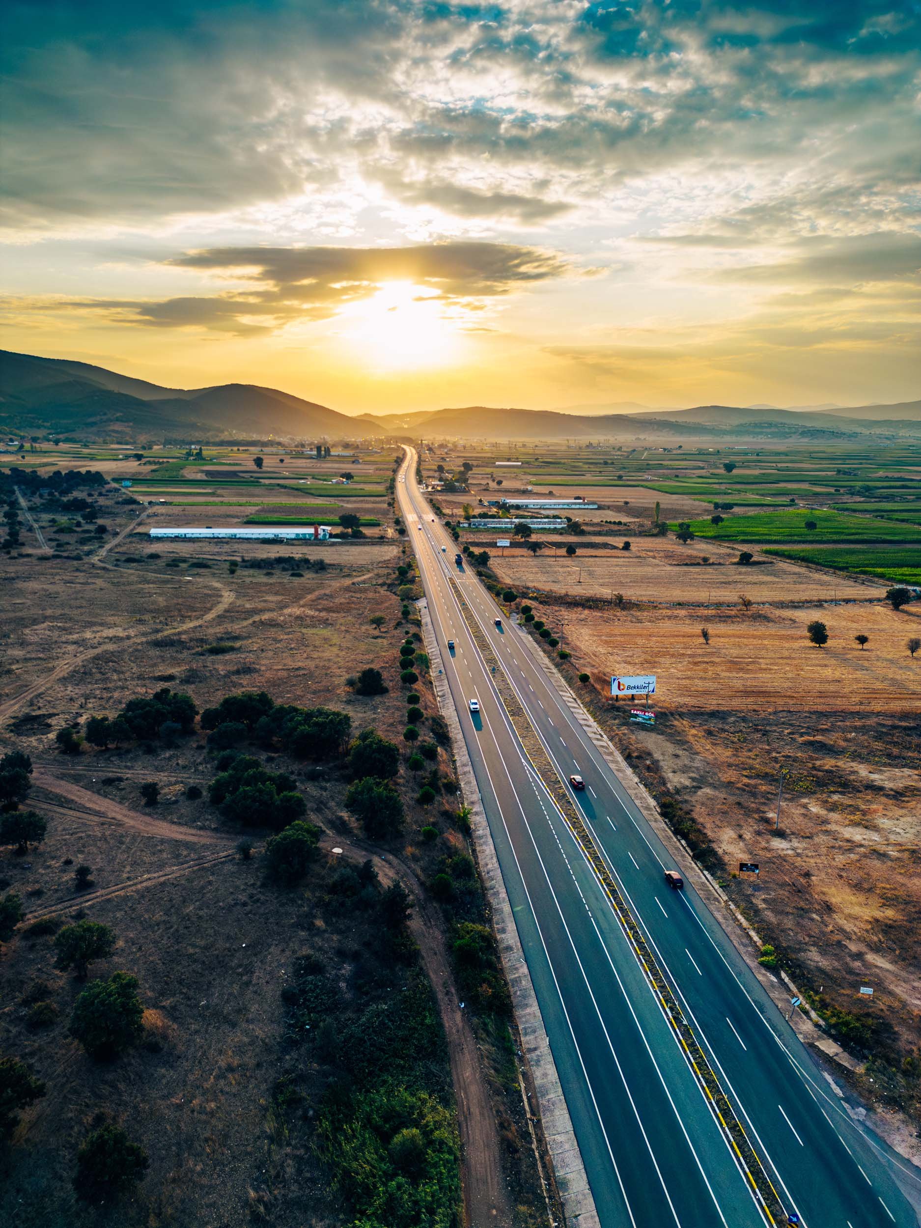 Aerial view of a highway extending through a rural landscape at sunset, with mountains in the background and cloudy sky overhead.