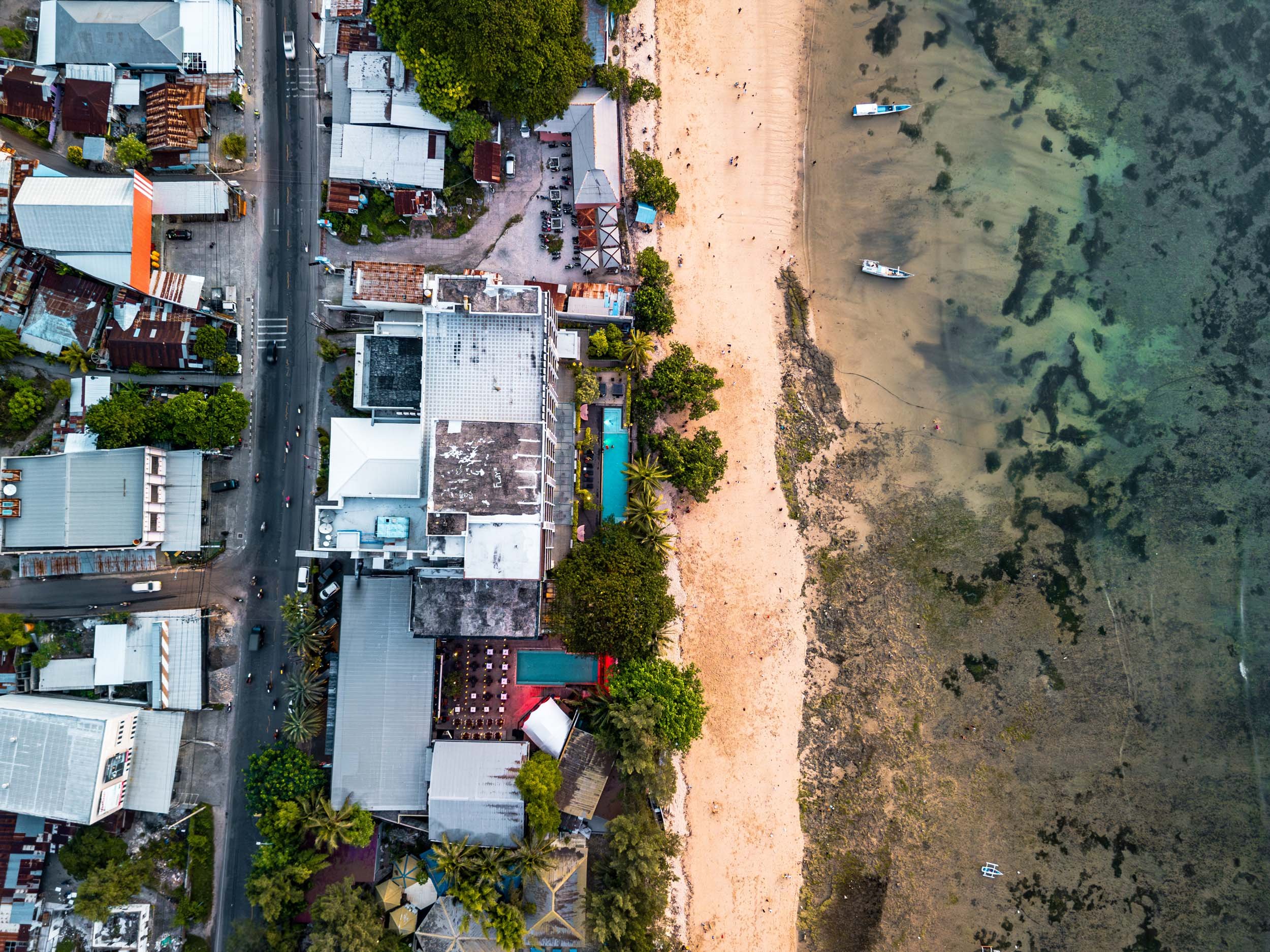 Aerial view of a coastal area showing a street lined with buildings, trees, and a beach with boats on the water.