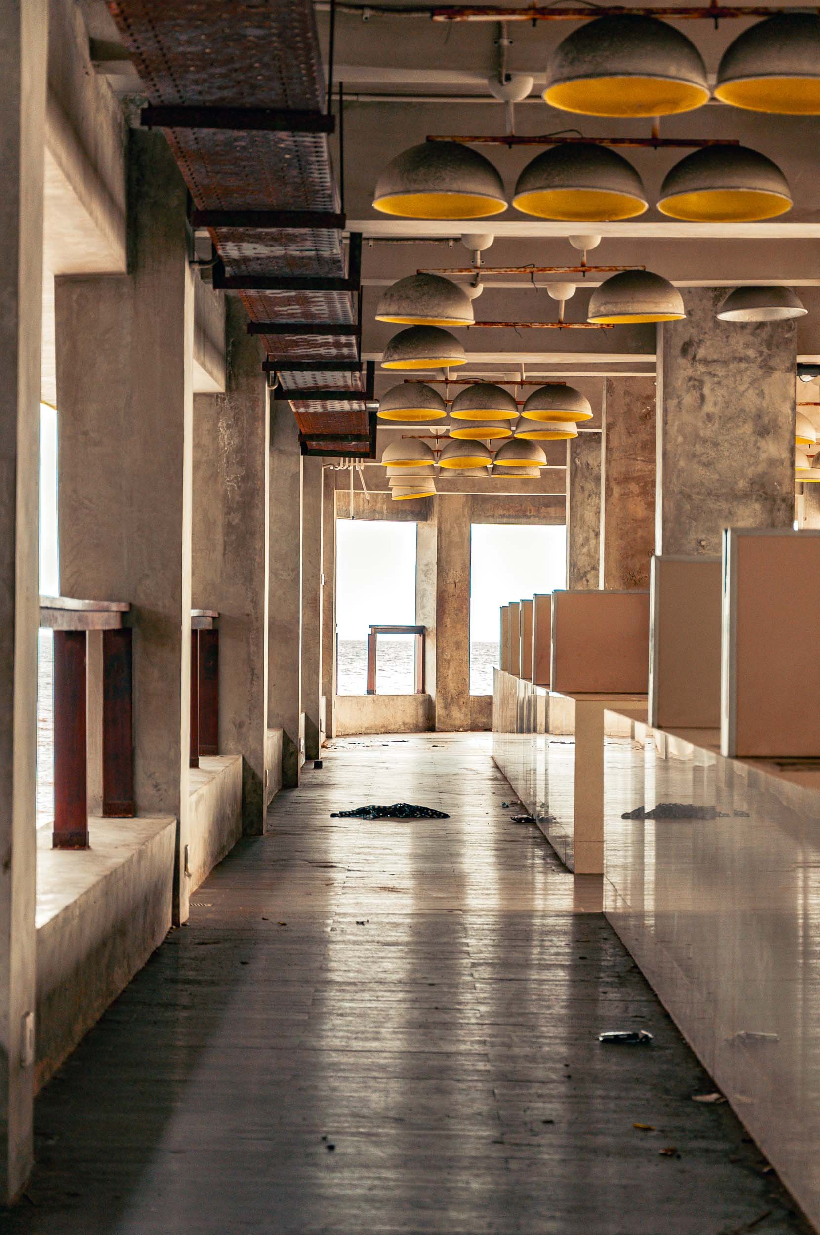 Empty restaurant with pendant lights hanging from the ceiling, large windows at the end showing an ocean view, and a wood floor with some debris scattered around.