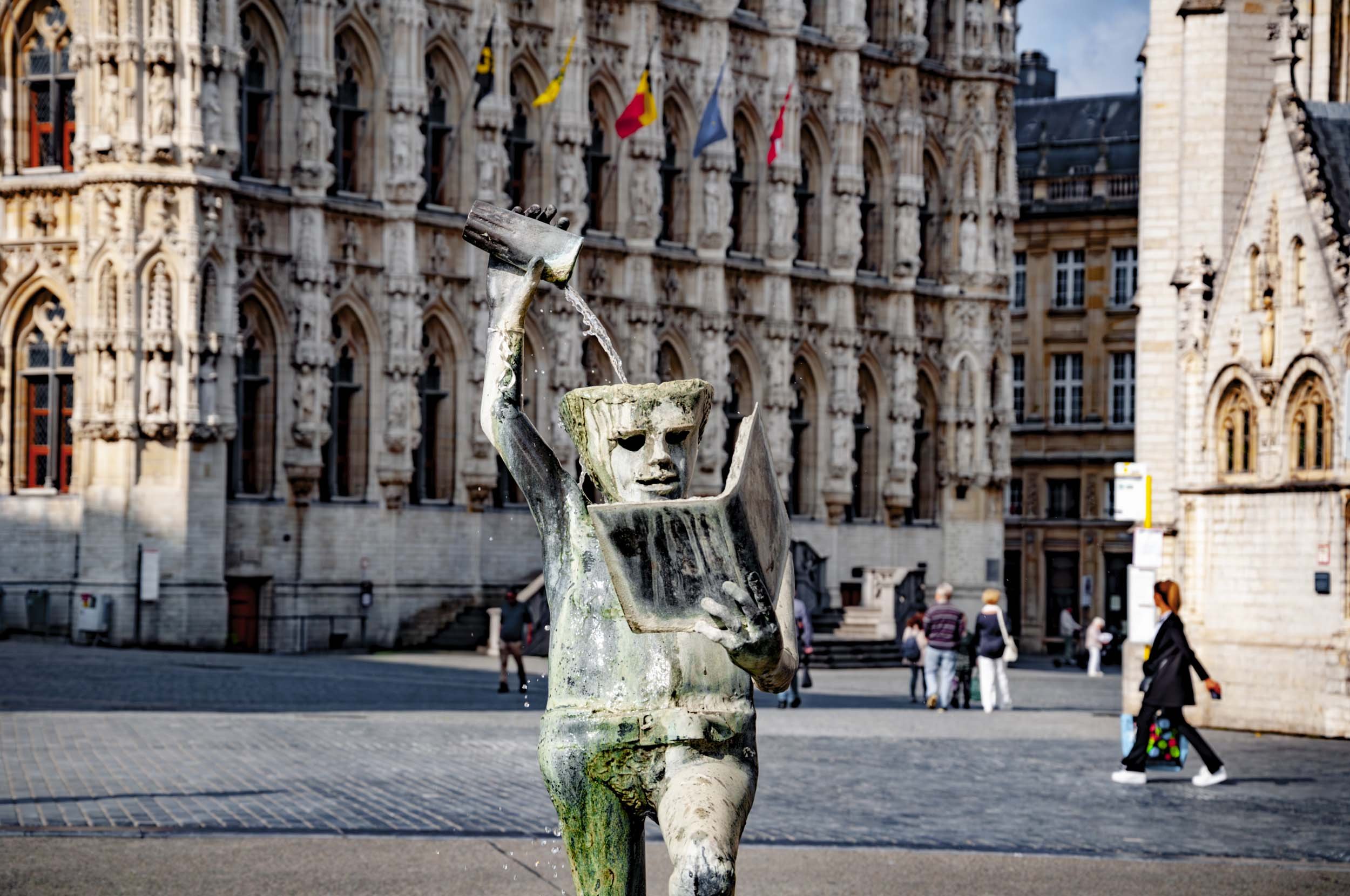 A weathered bronze sculpture of a child reading a book while holding a watering can. Water pours from the can onto the book. The sculpture is located outdoors in a city square with historic stone buildings and pedestrians in the background.
