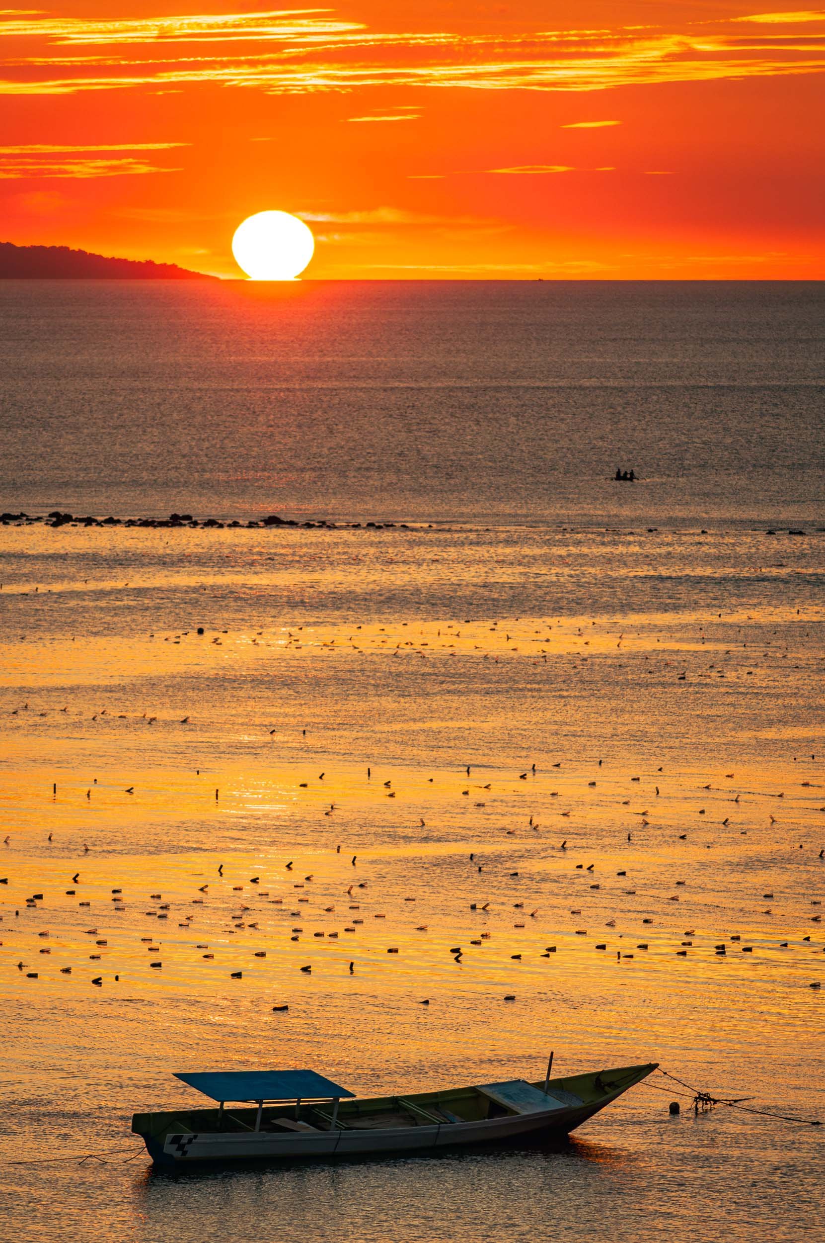 A sunset over a calm ocean with orange and yellow hues. A boat floats near the shore, and birds are visible on the water's surface. There are dozens of plastic bottles in the water.
