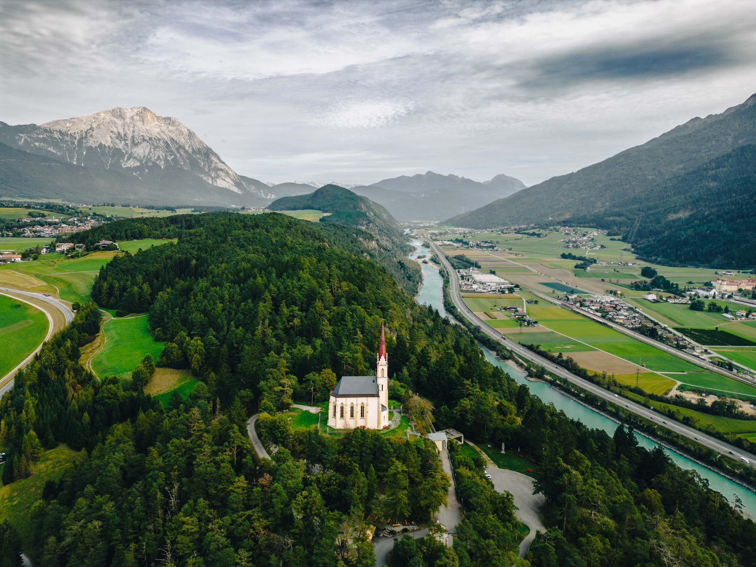 Aerial view of a church on a forested hill overlooking a river, surrounded by fields, Dolomite Alps at the background