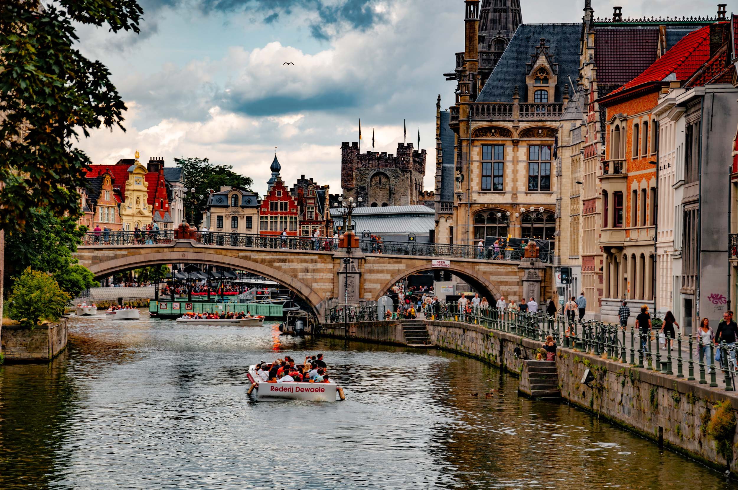 Scenic view of a canal in Ghent, Belgium, with a boat filled with tourists, historic medieval buildings, a stone bridge, and a castle tower in the background under a partly cloudy sky.