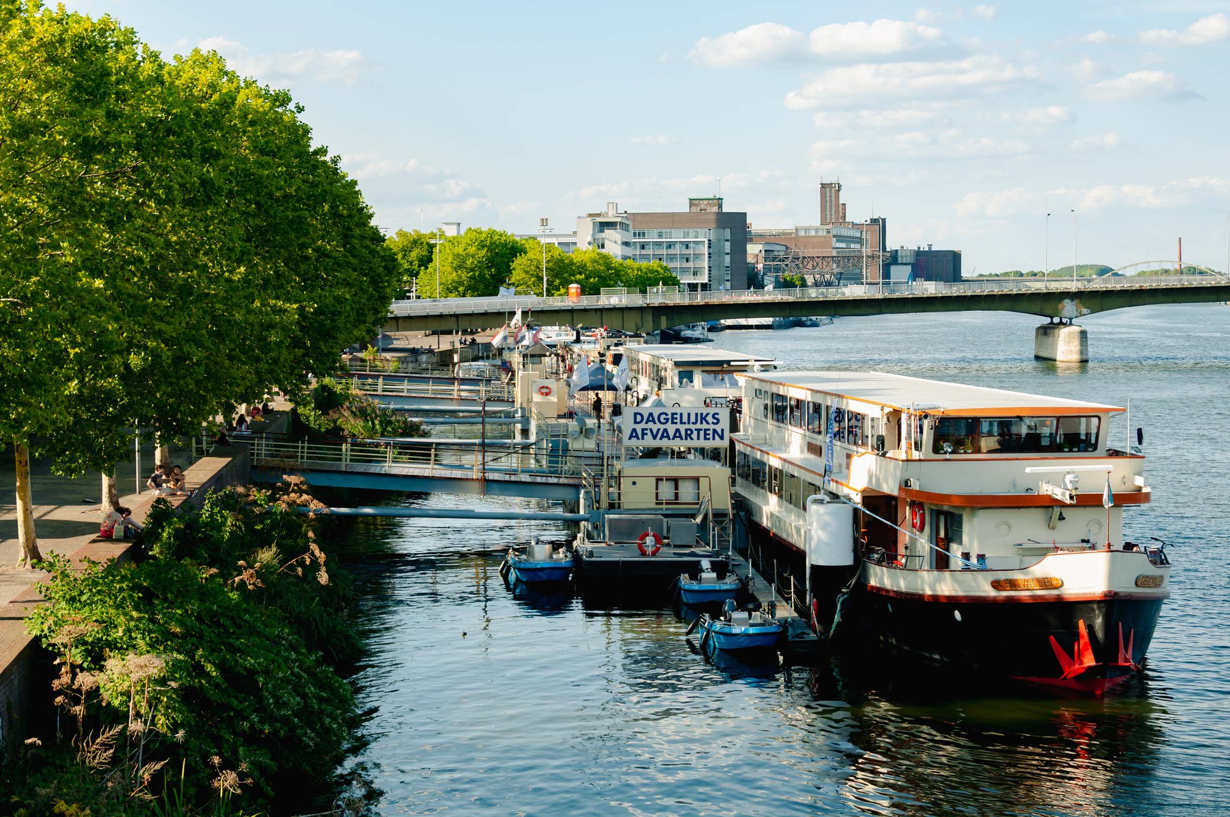 A river scene with a large white boat docked at a pier, small boats tied to it. The river has a bridge overhead, with a cityscape in the background under a partly cloudy sky. People are sitting on the riverside walkway shaded by green trees.