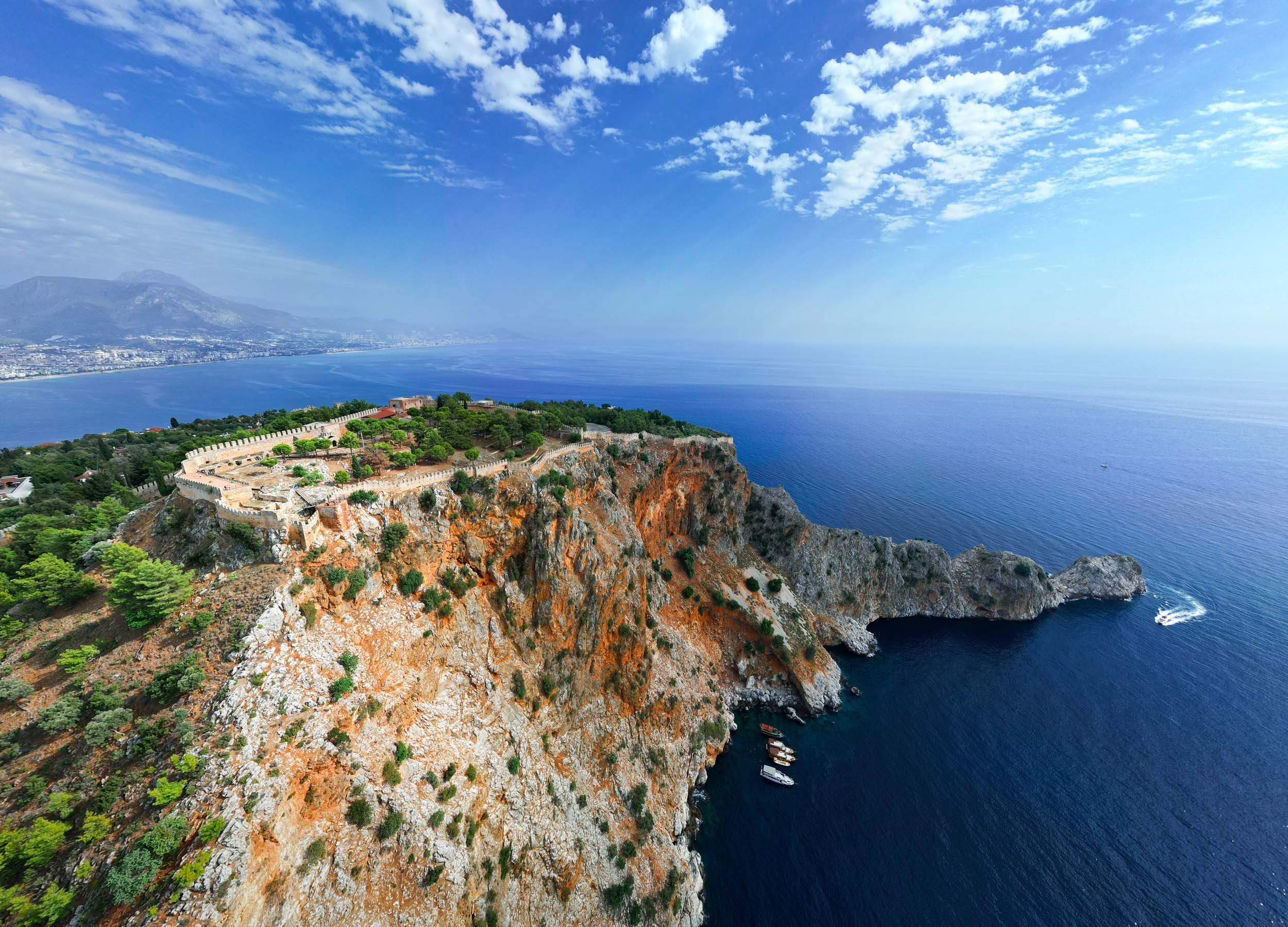 Alanya aerial view of a coastal fortress on a rocky cliff with boats in the water below and a city in the distance under a partly cloudy sky.