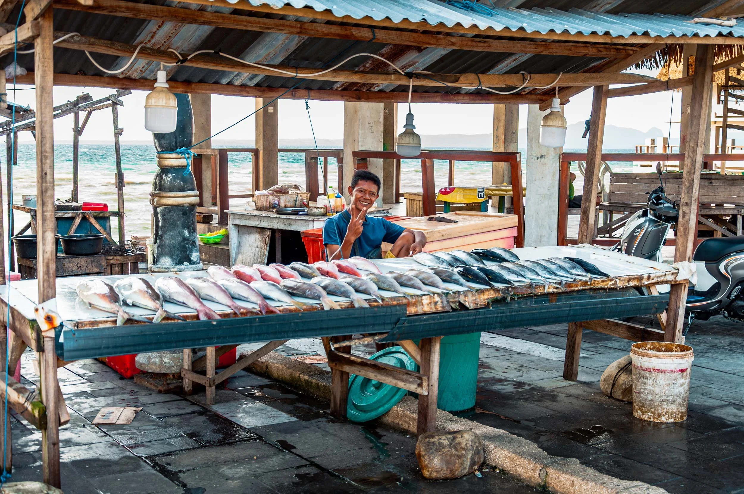 A smiling man at a seaside fish market, selling various types of fresh fish laid out on a long wooden table under a rustic shelter.