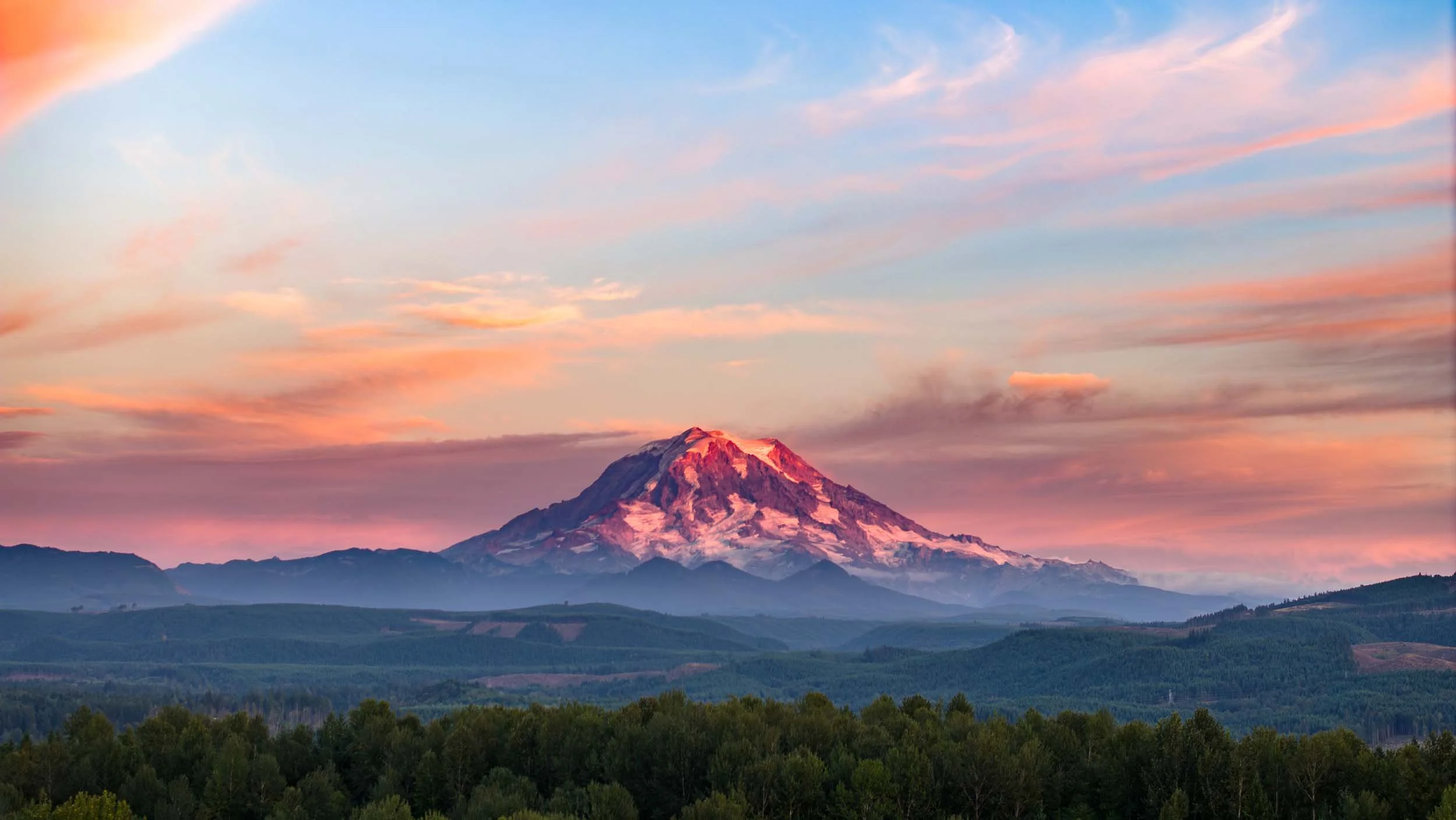 Snow-capped mountain at sunset with colorful sky and green forest in the foreground.