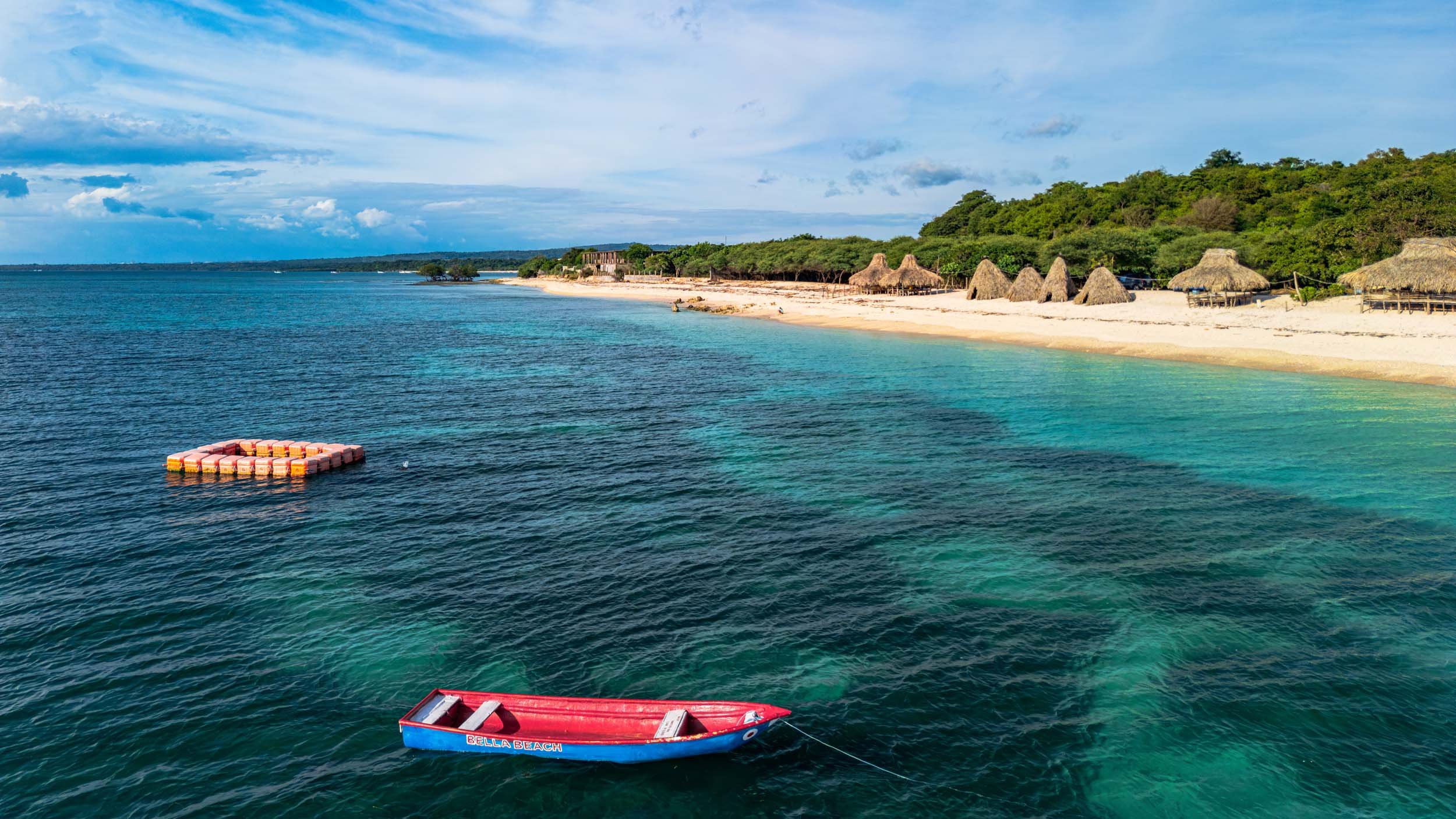 Beach with clear blue water, thatched umbrellas on the sandy shore, and a small boat named 'Bella Beach' floating on the water.