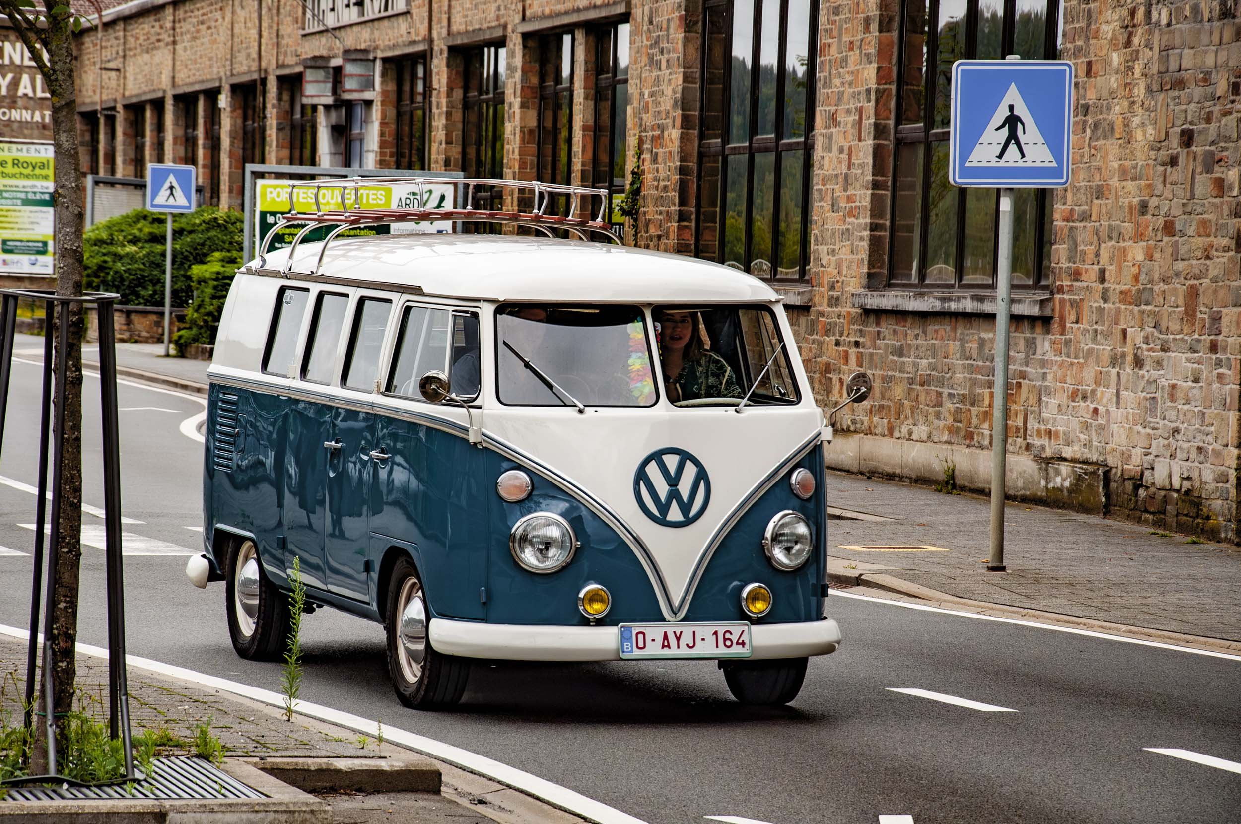 A vintage blue and white Volkswagen Type 2 van driving on a city street during daytime with pedestrian crossing signs visible.