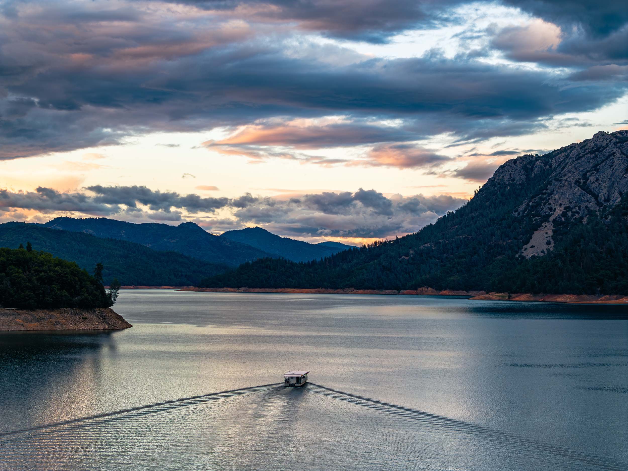 A boat cruising on a large lake surrounded by mountains and forest under a partly cloudy sky during sunset.