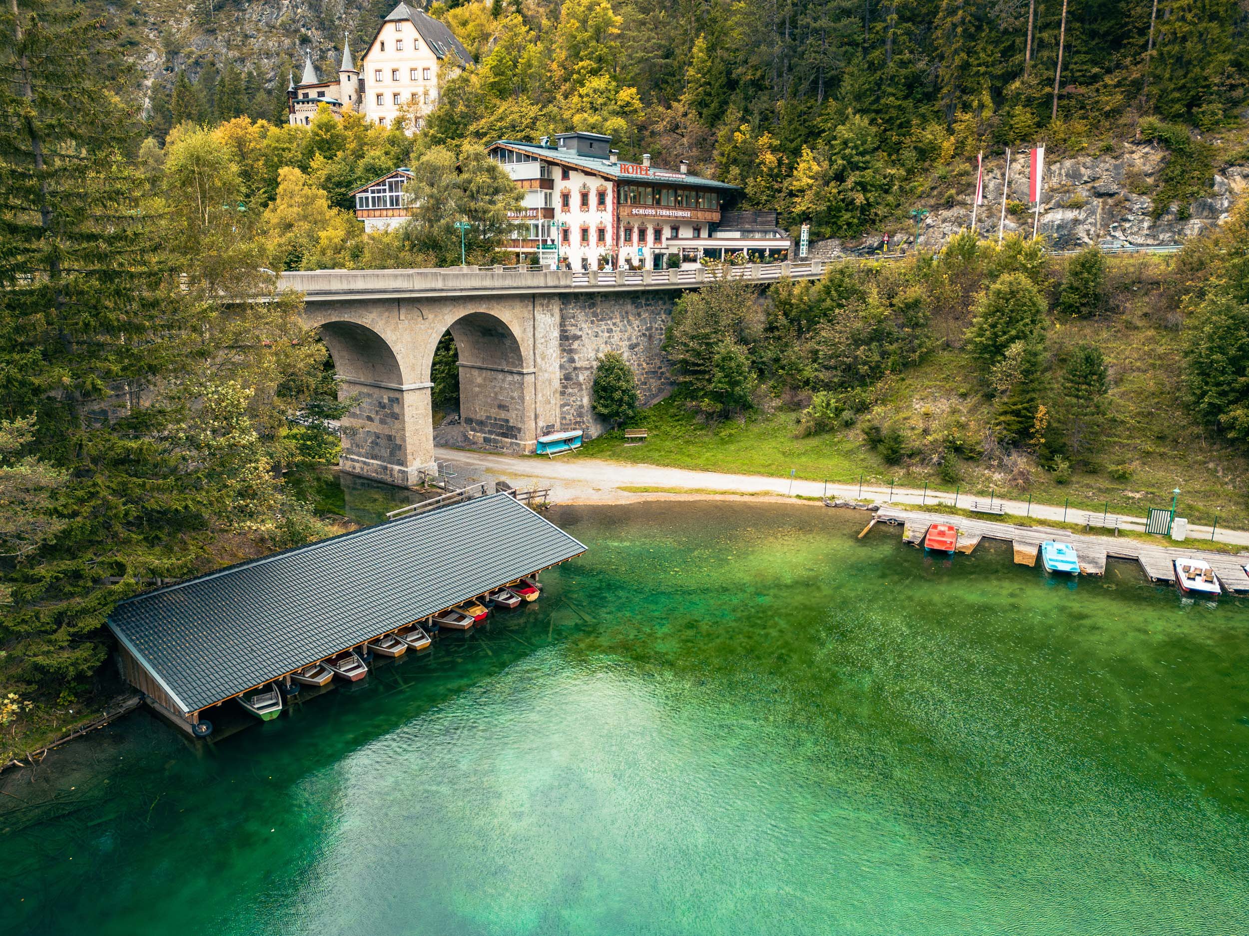 A scenic view of a lake with a covered boat dock housing multiple boats, surrounded by green trees and hillside buildings, including a hotel and castle in the background.