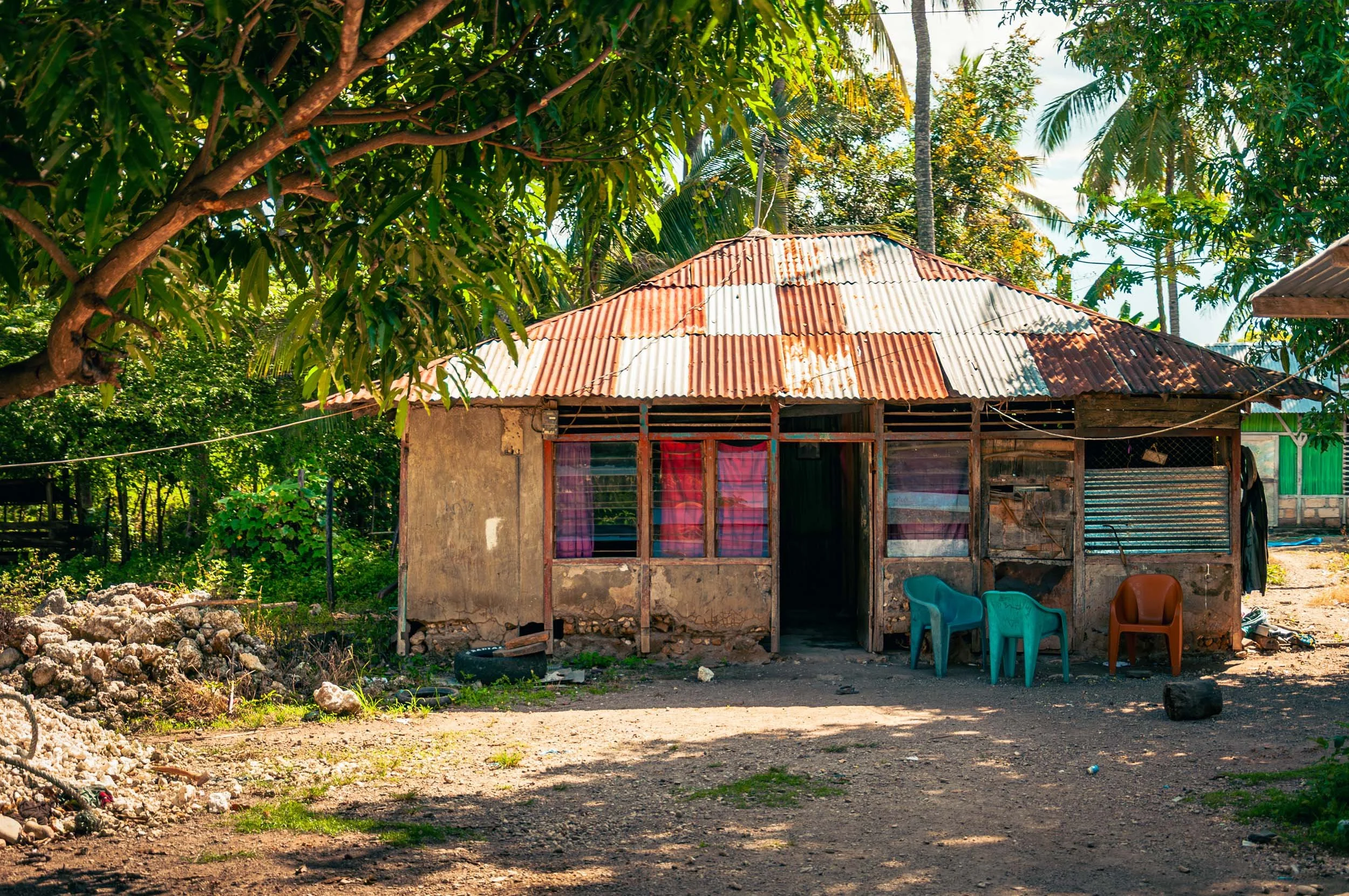 A small, weathered house with a patchwork metal roof, pink curtains, and a dirt yard with plastic chairs outside, surrounded by lush green trees.