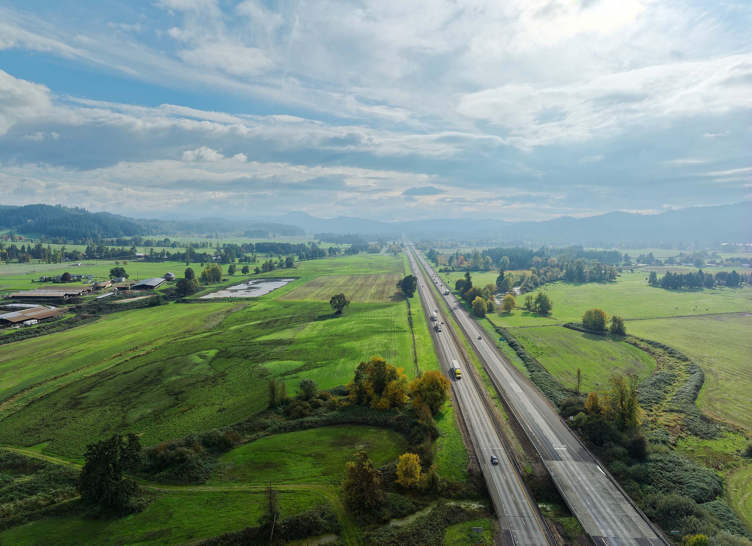 Aerial view of a rural landscape with fields, a highway with cars, trees, and distant mountains under a partly cloudy sky.