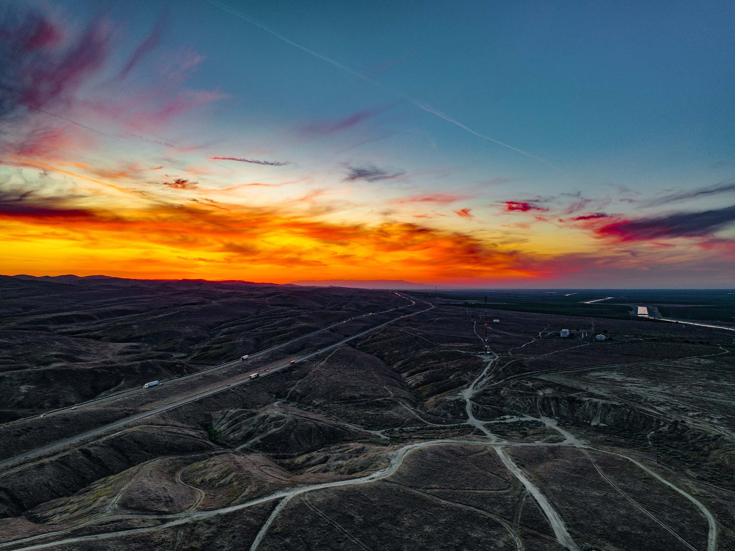 A landscape scene at sunset showing a hilly terrain with dirt roads, a highway with trucks, and utility poles, with the sky filled with colorful clouds and contrails.