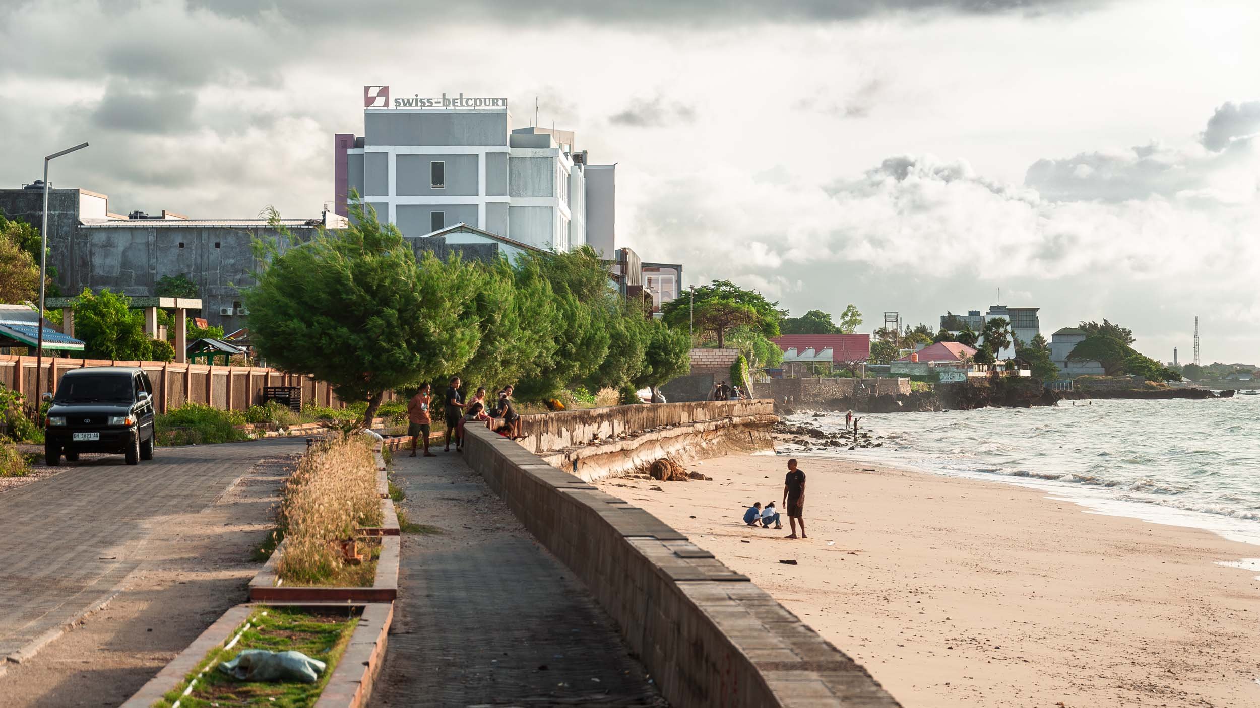 Beachside promenade with people walking and sitting, sandy beach with a person walking and two children playing, ocean waves, buildings including a high-rise with Swiss-Belcourt sign, trees, cloudy sky.