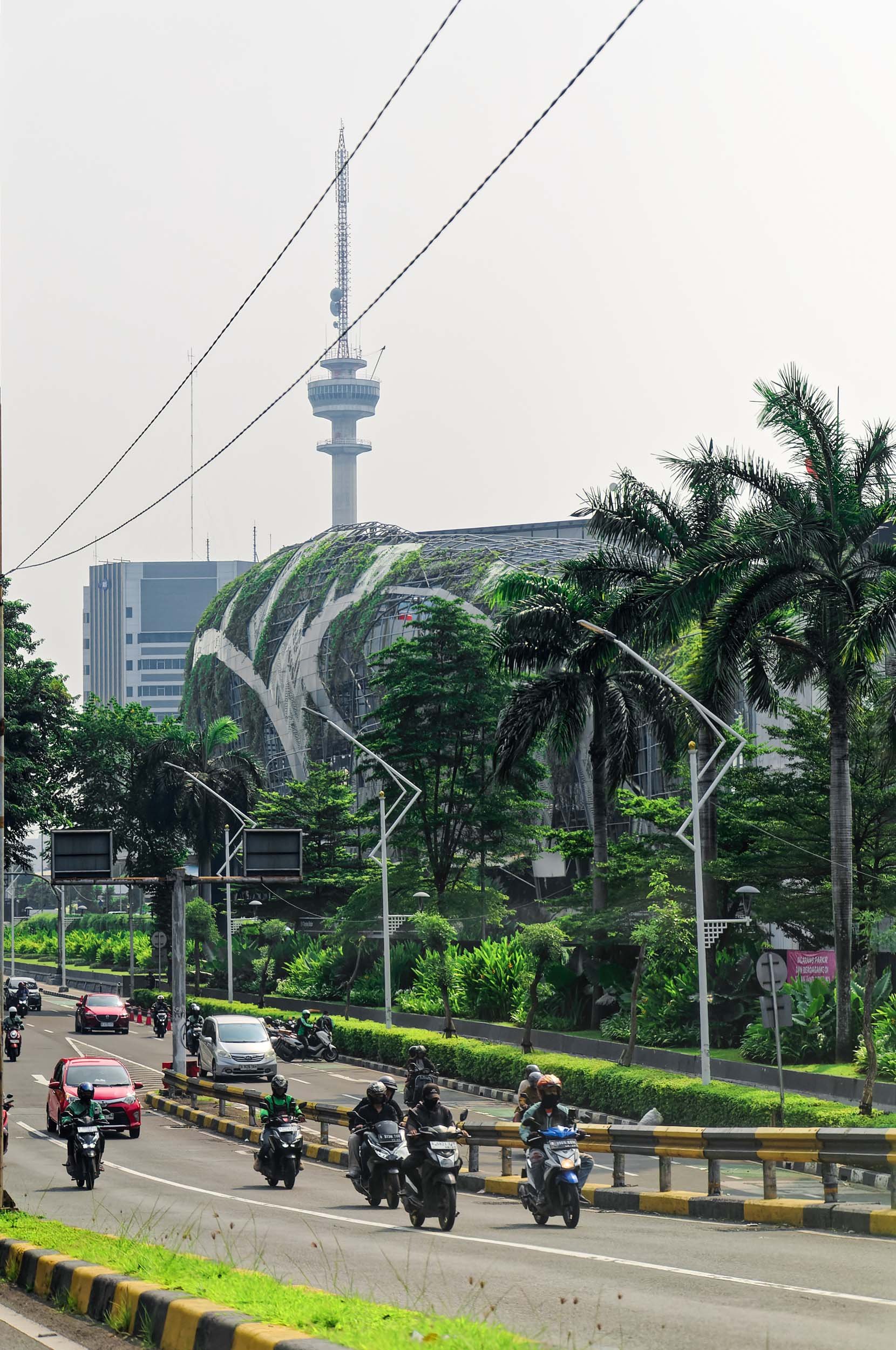 A busy street with motorcycles, cars, and lush greenery, with a modern building in the background featuring a glass roof and a tall telecommunications tower.