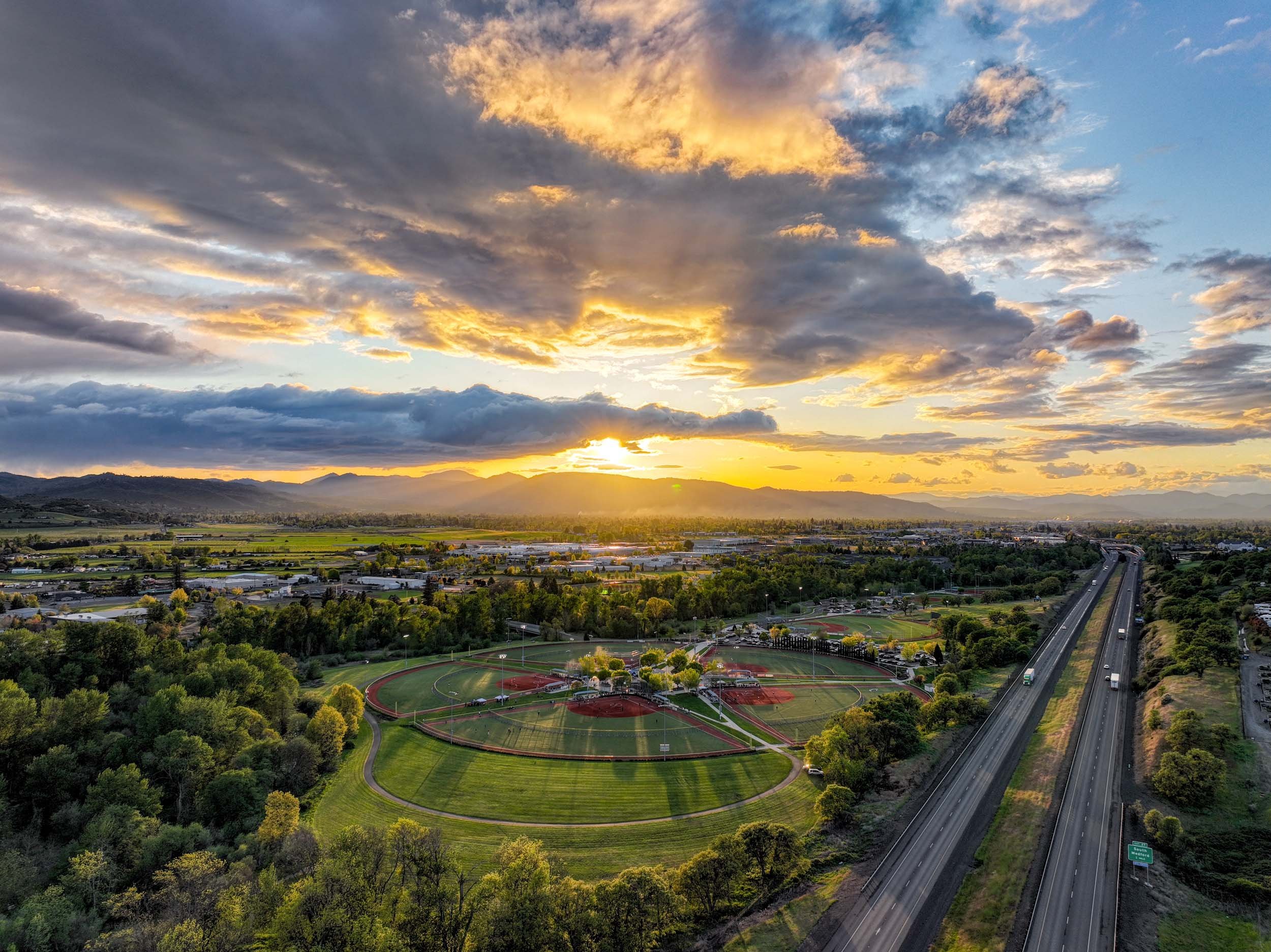 Aerial view of a park with baseball fields, a running track, and trees, during sunset with mountains and a highway in the background.