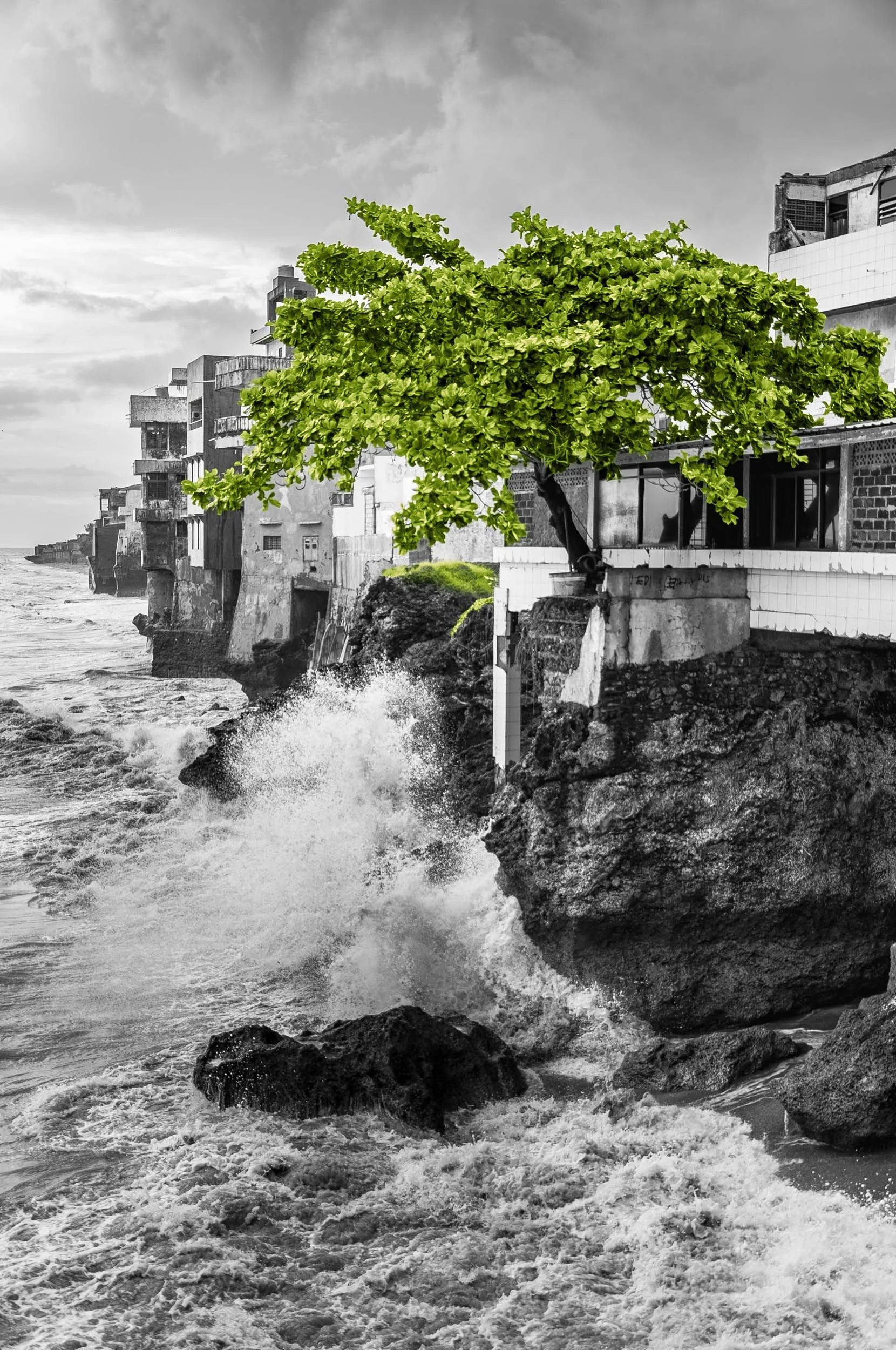 A photograph of a coastal scene showing waves crashing against rocks below a white building with a tree growing on the edge, with the background of a cloudy sky and multiple similar buildings on the cliff.