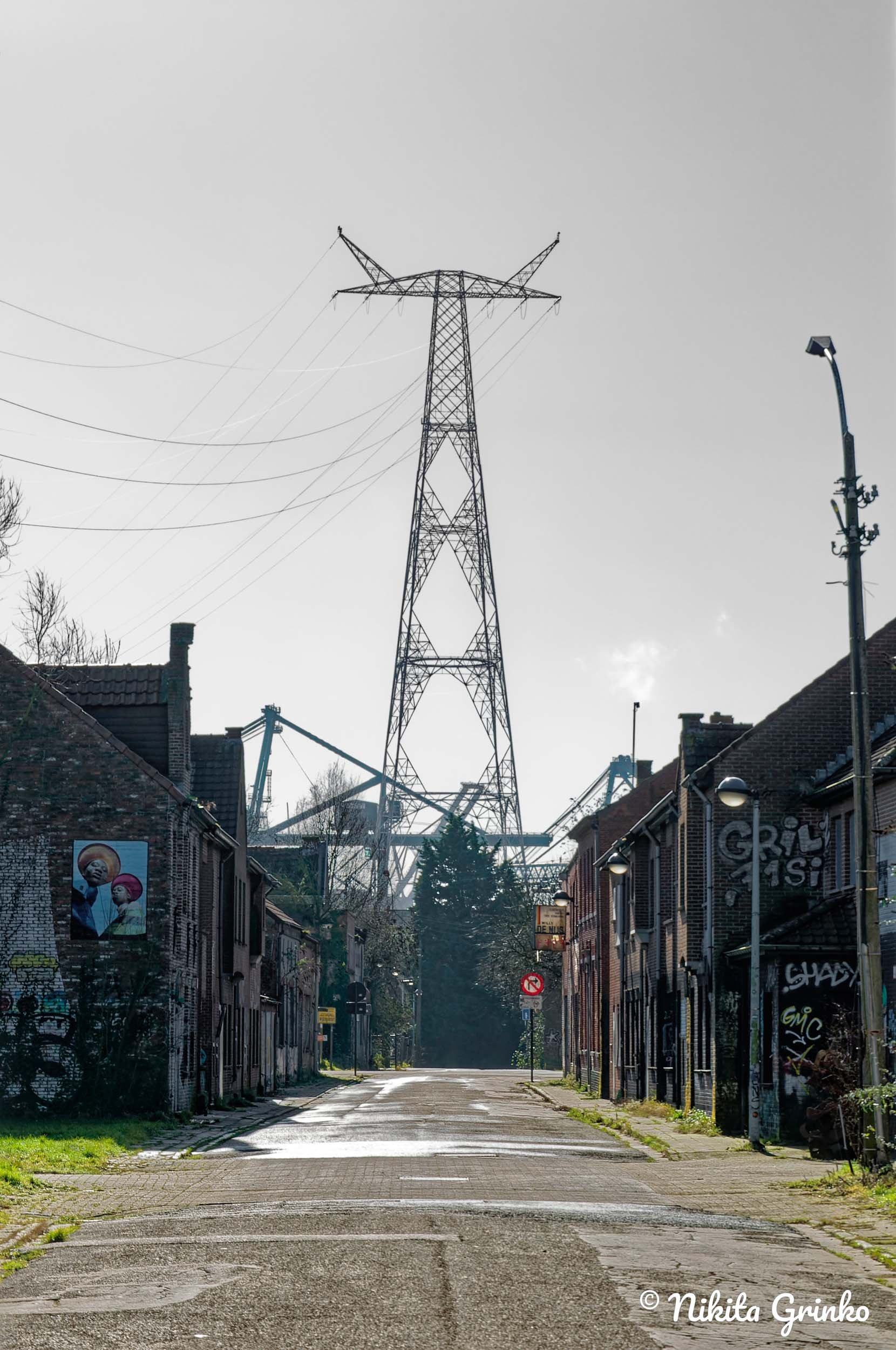 An urban alleyway with low-rise brick buildings on either side. Graffiti is visible on the walls, and streetlights illuminate the scene. A large power transmission tower is prominent in the background, towering over the buildings and crossing wires.