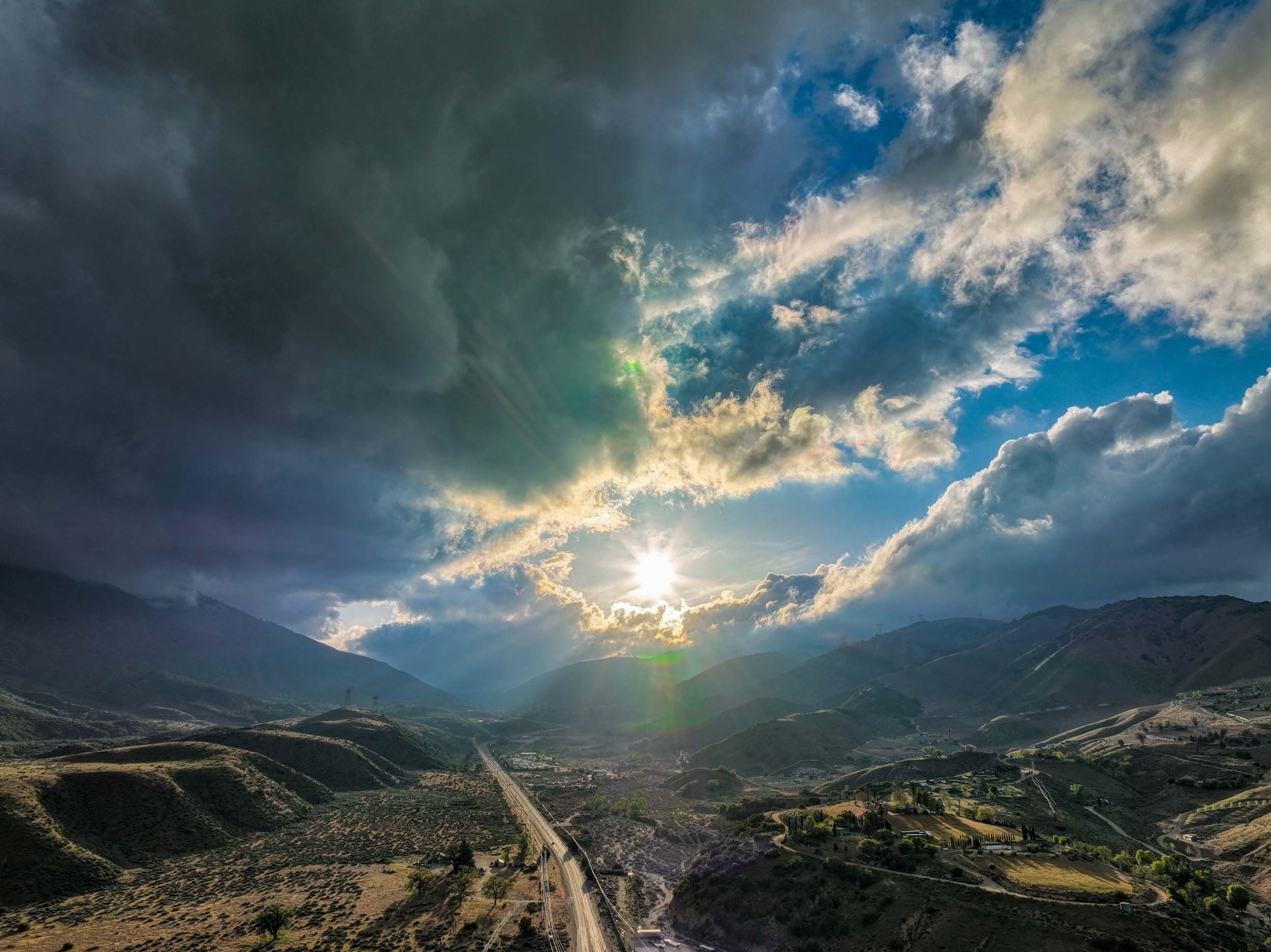 Sun shining through clouds over rolling hills and mountains in a rural landscape.