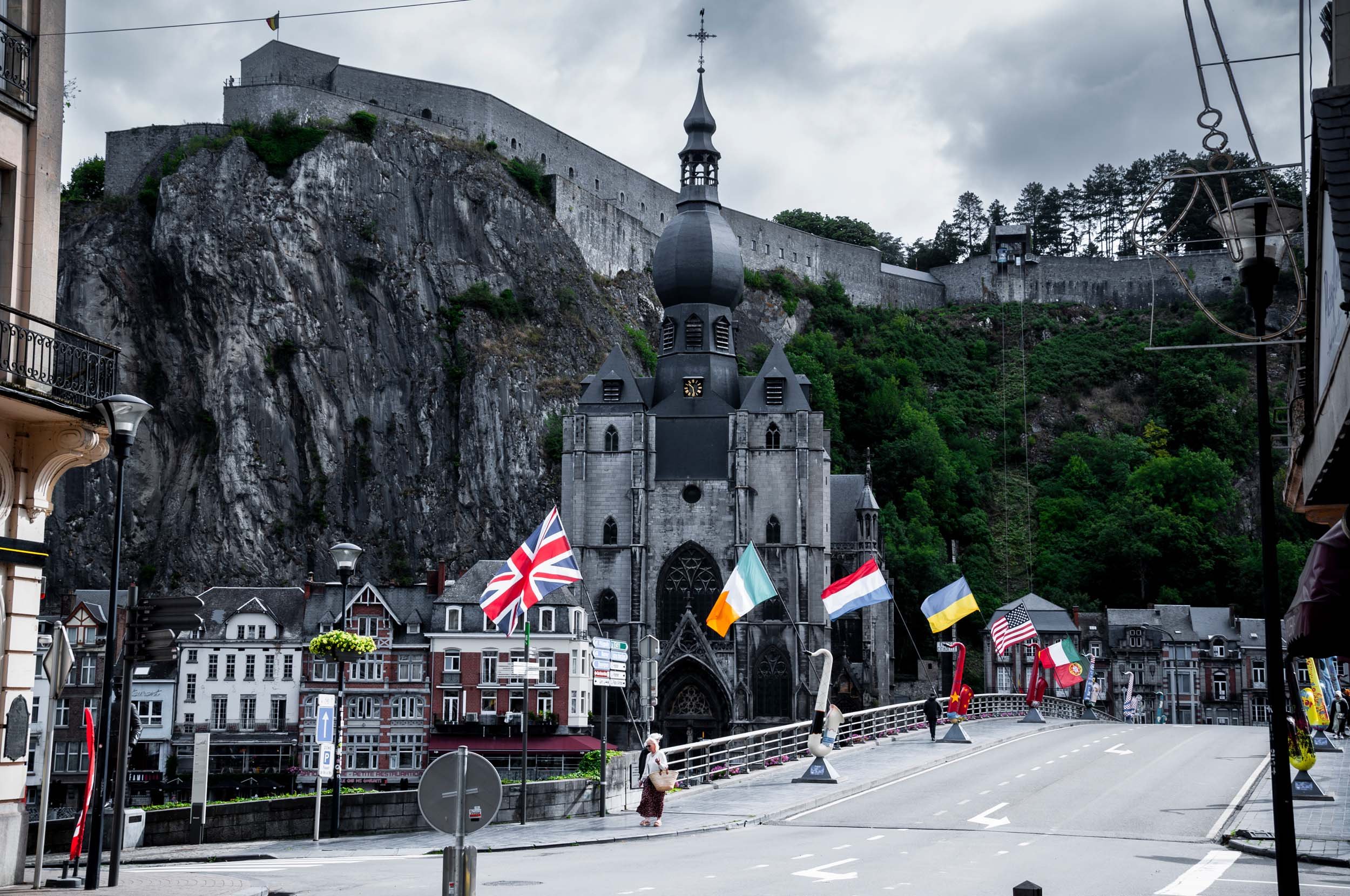 A street scene with a church in the foreground, flags including the UK, Irish, Dutch, Belgian, American, Ukrainian and Portuguese flags, and a large castle on a rocky hill in the background, with a cloudy sky overhead.