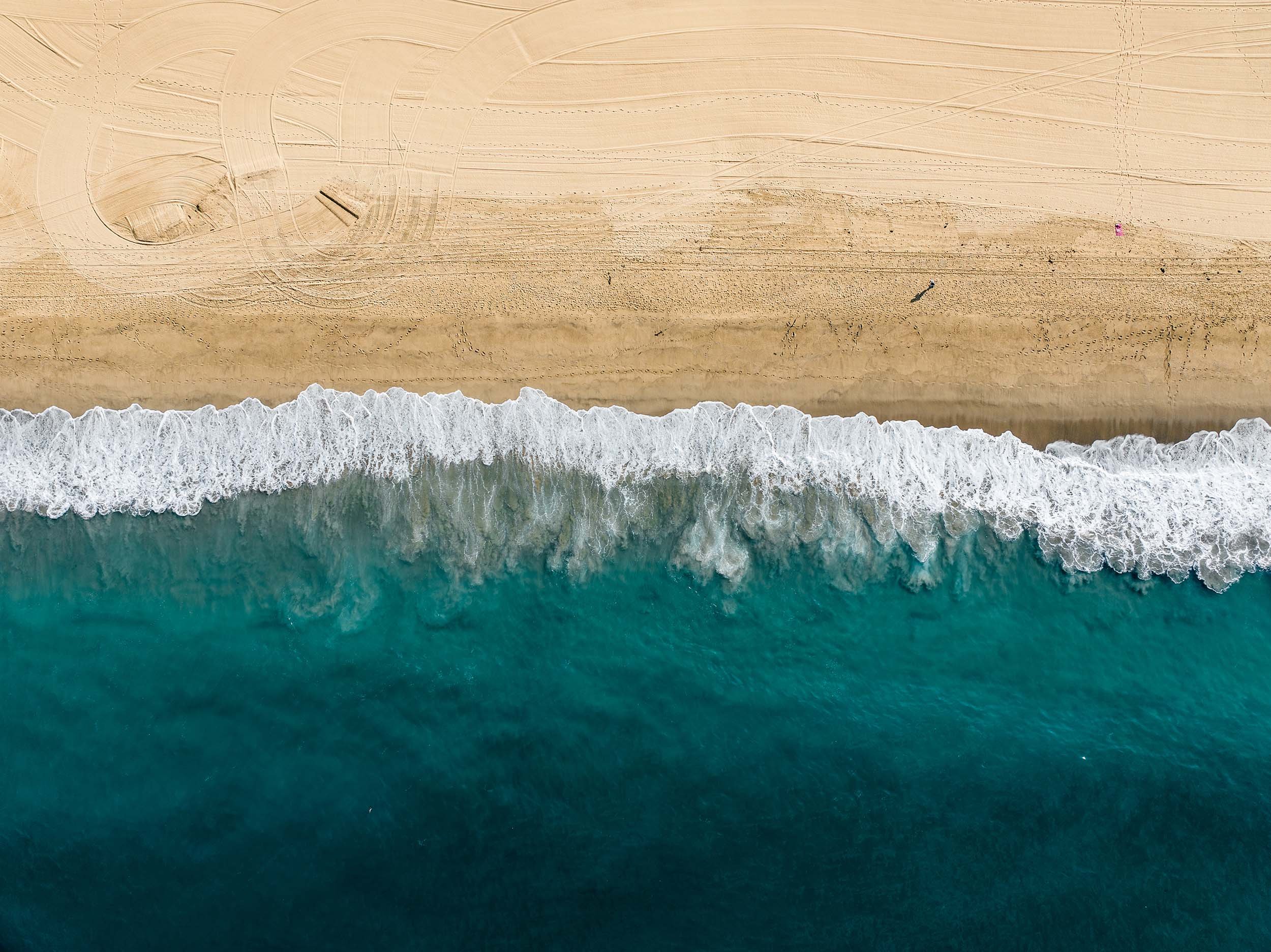 Aerial view of a sandy beach with waves crashing onto the shore, showing tire tracks and footprints on the sand.
