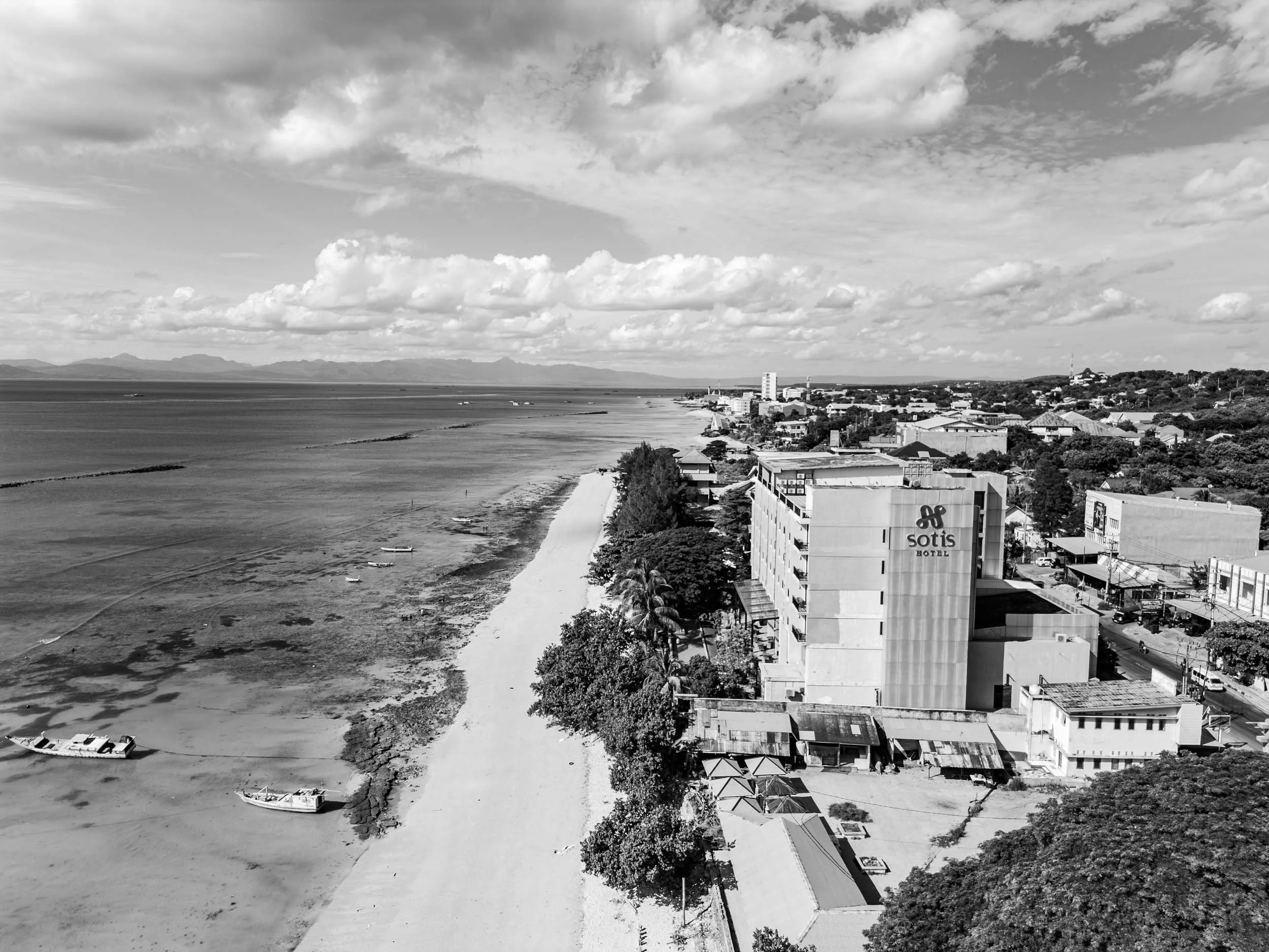 Black and white aerial view of a coastal town with a beach, boats, and a hotel labeled 'Sotis Hotel' near the shoreline.