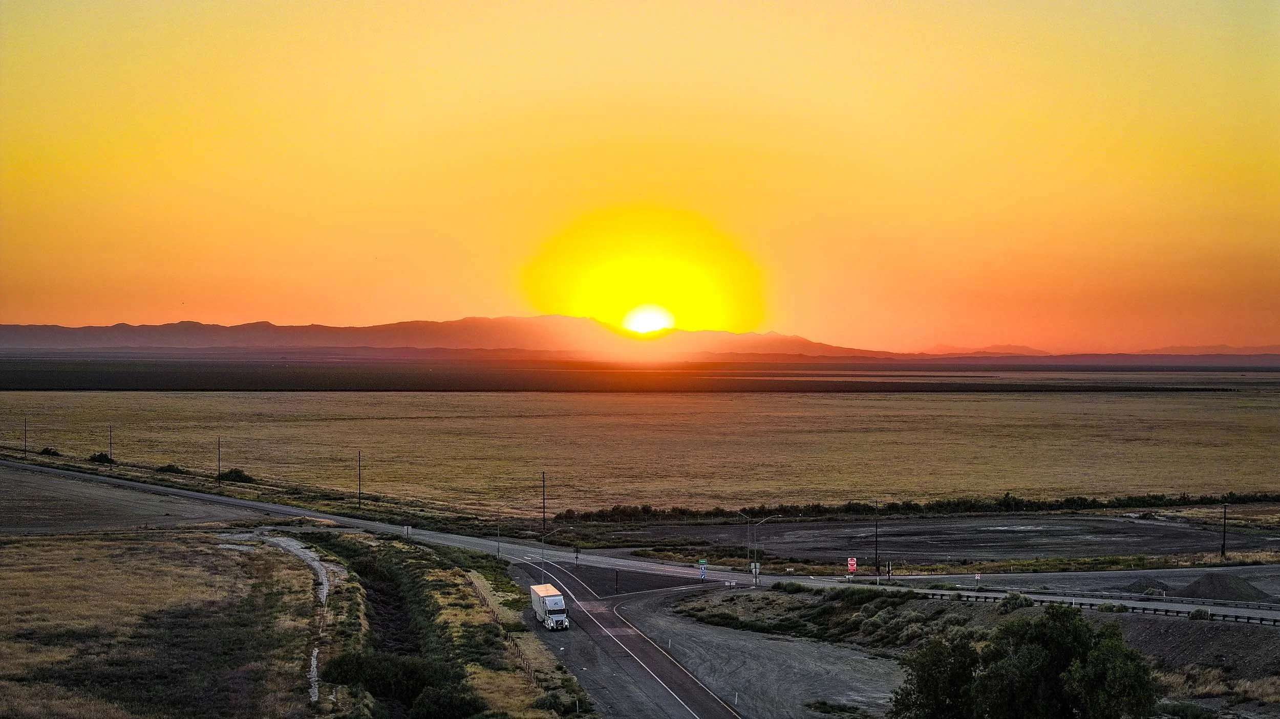 Sunset over a flat, rural landscape with a truck on a winding road in the foreground.