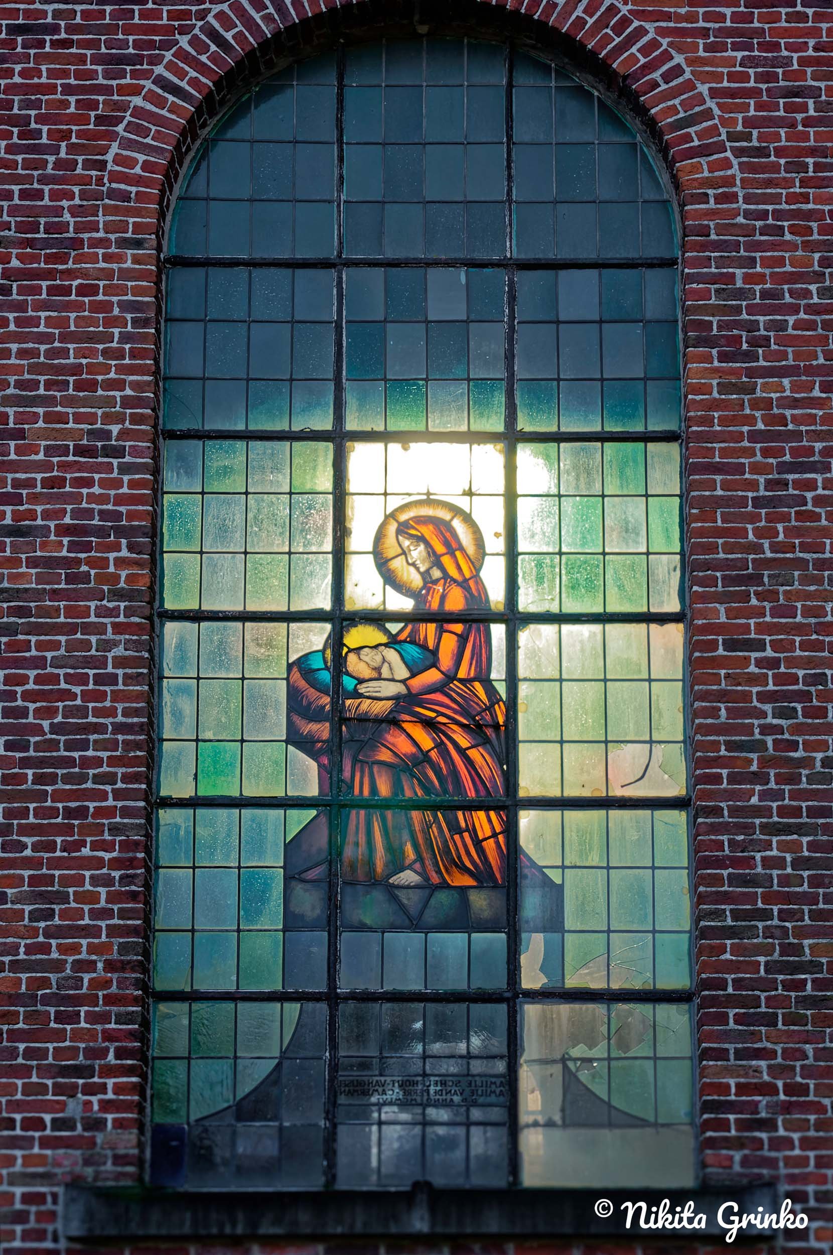 Stained glass window depicting the Virgin Mary holding baby Jesus, set in a brick wall.